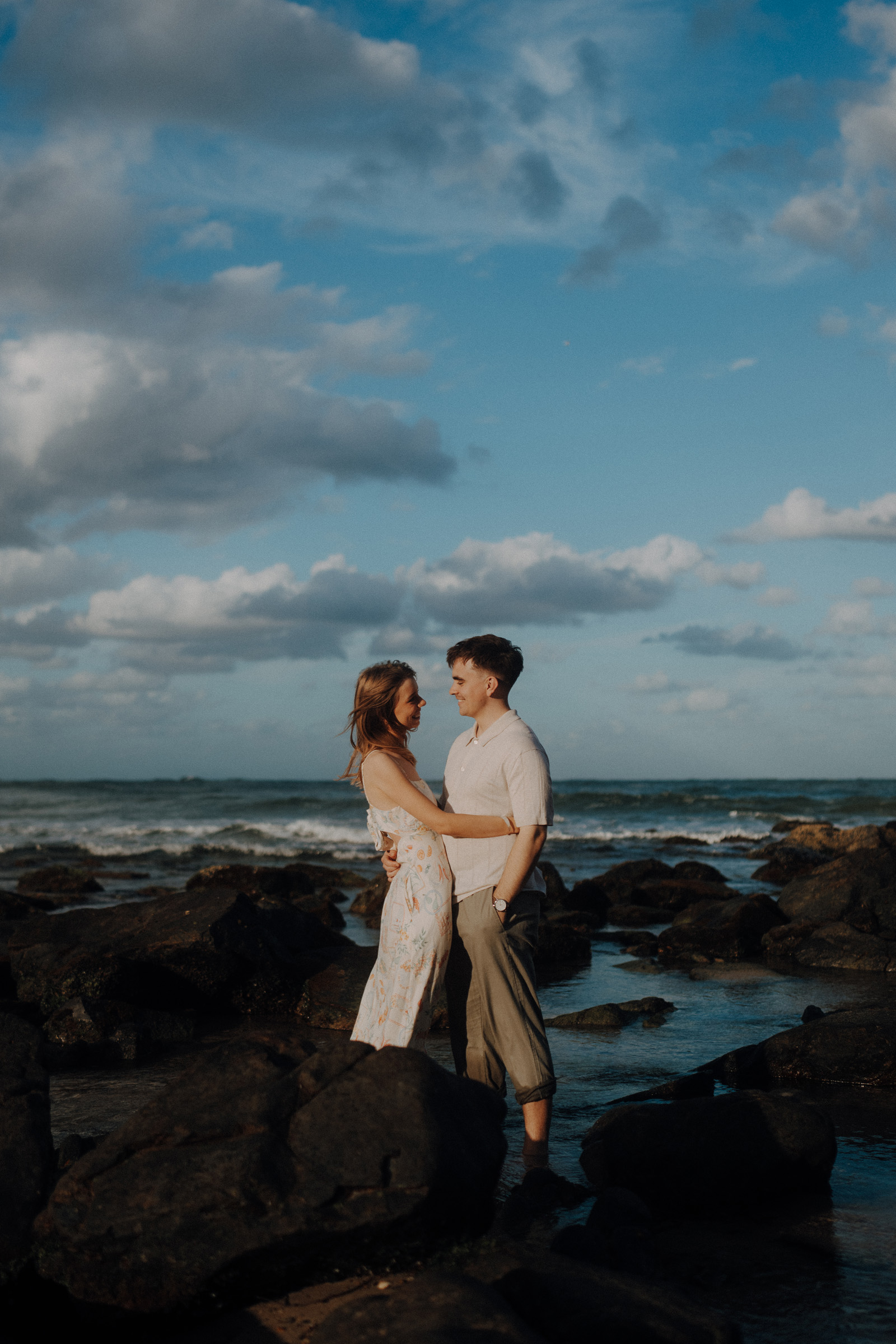 A couple stands embracing on rocks by the ocean under a partly cloudy sky.