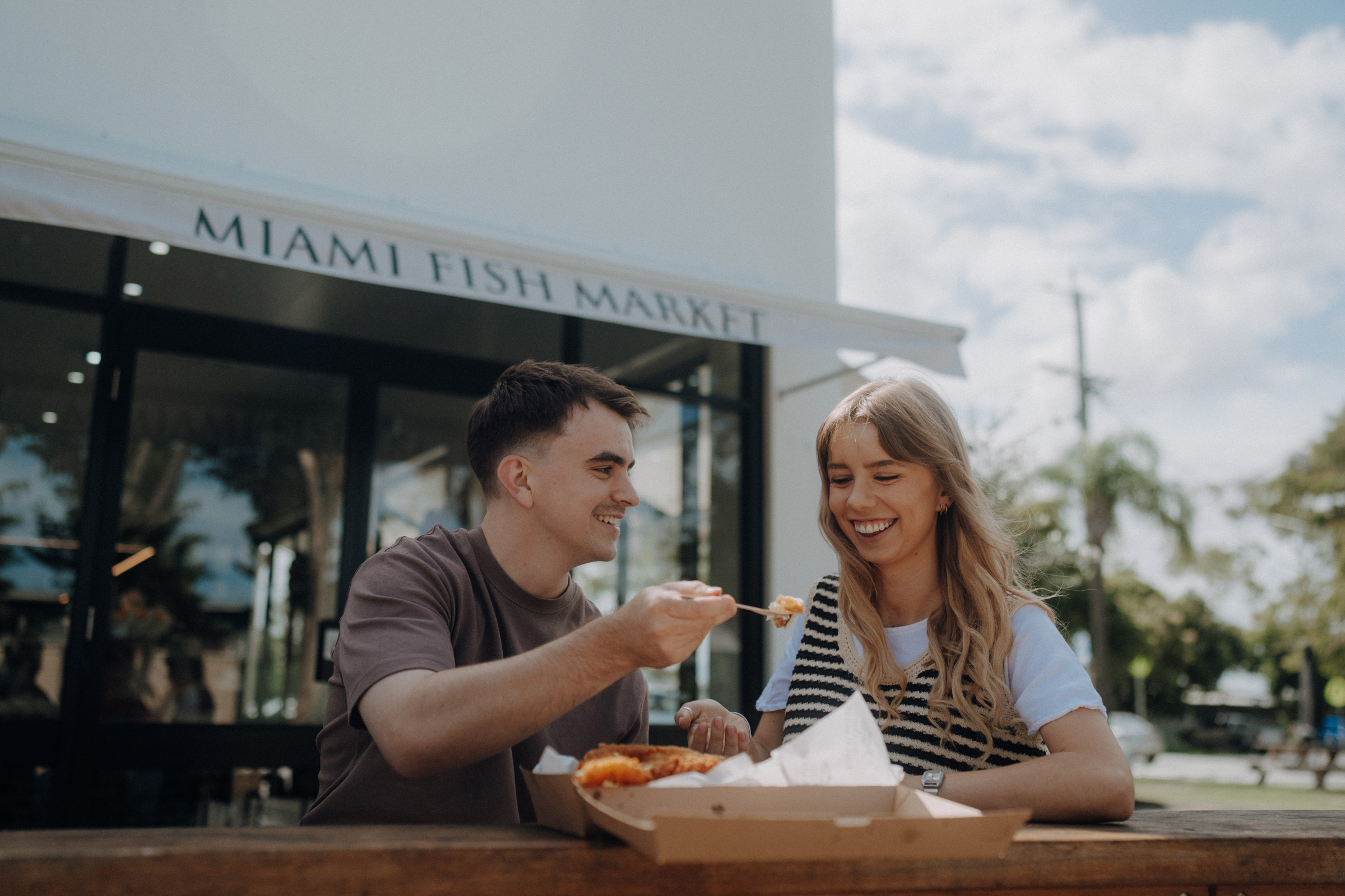 A man and woman sharing a meal at an outdoor table in front of Miami Fish Market. The man offers food to the woman, who is smiling.