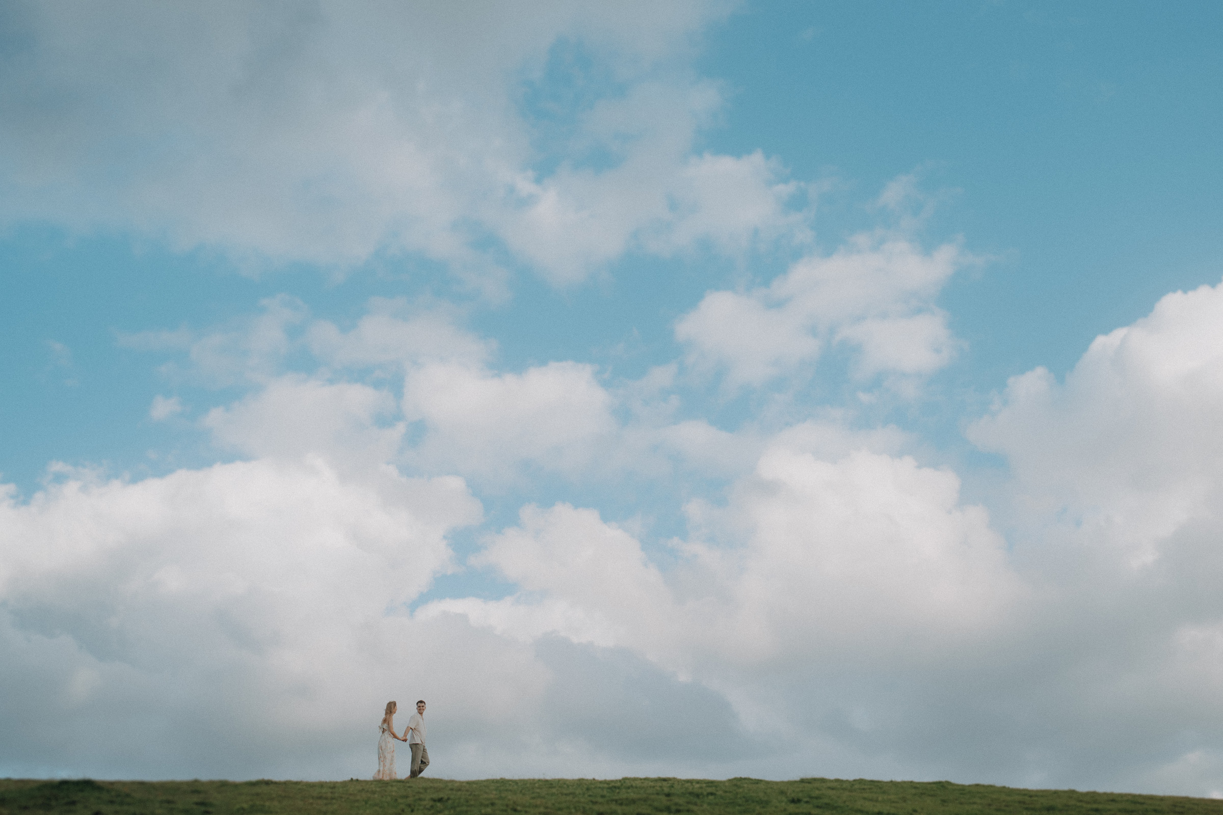 A couple holding hands walks on a grassy hill under a bright blue sky with scattered clouds.