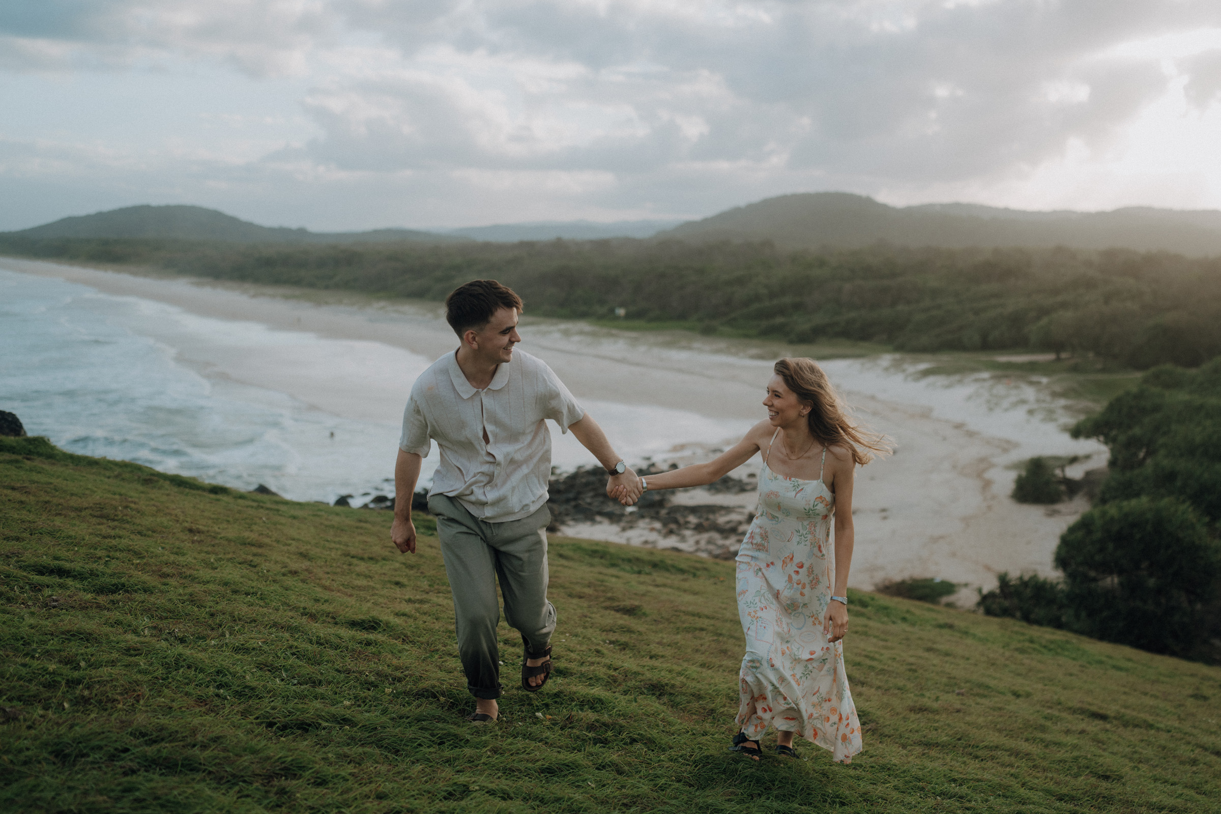 A man and woman hold hands while walking up a grassy hill by a beach, with the ocean and hills in the background under a cloudy sky.