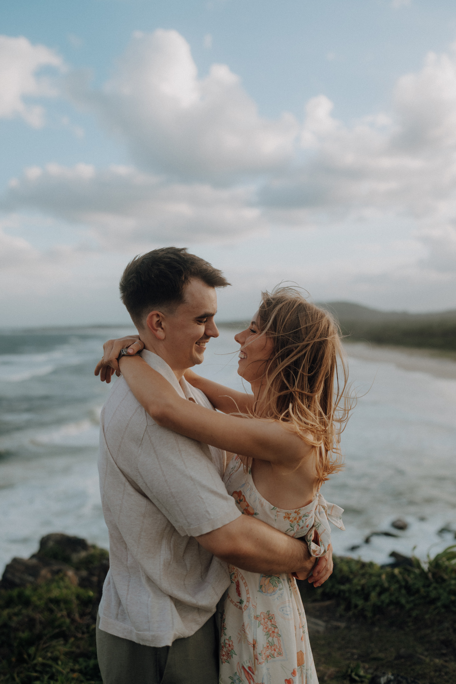 A couple embraces and smiles at each other by the seaside, with waves and a cloudy sky in the background.