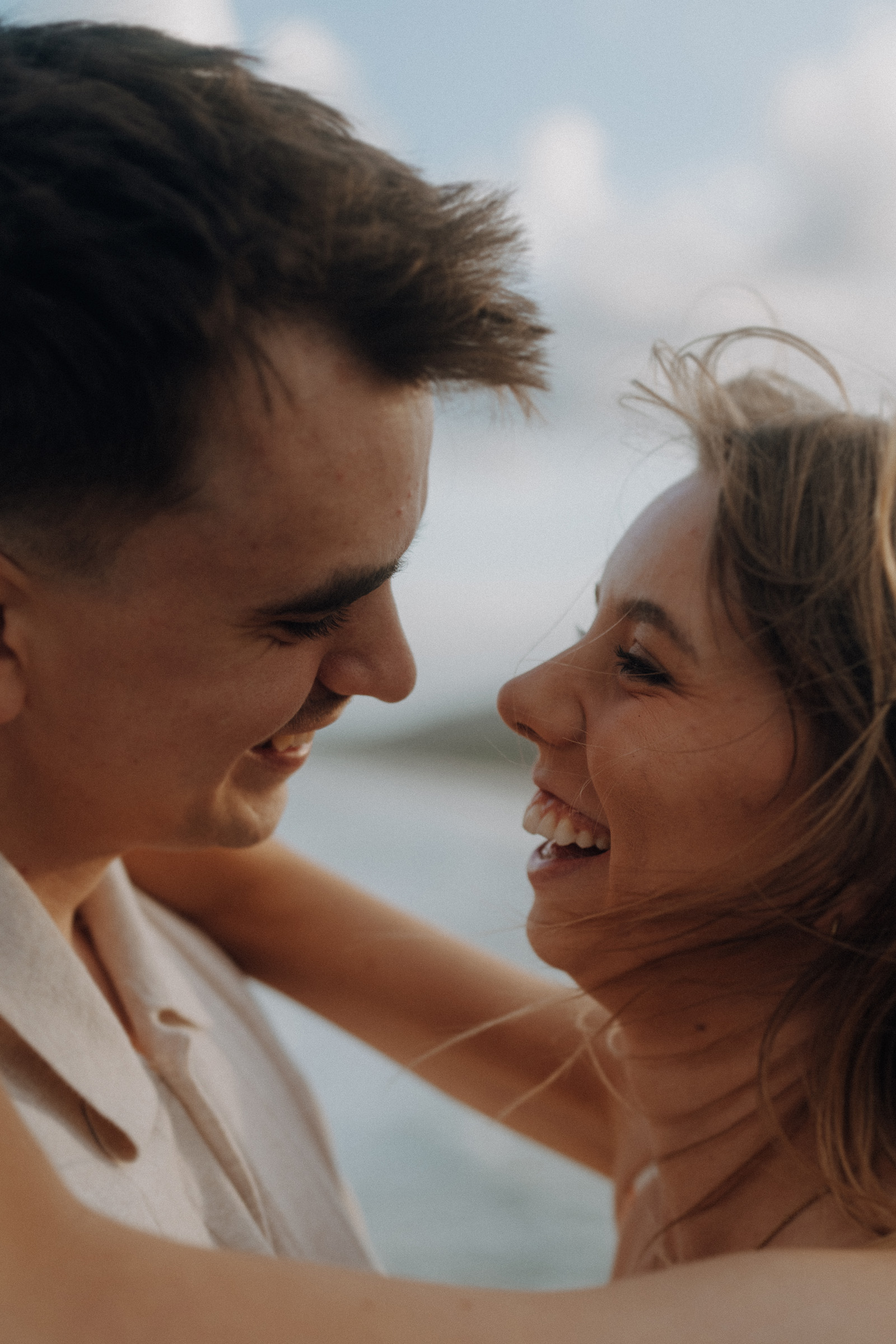 A couple smiling at each other closely, with a blurred background of the sea and sky.