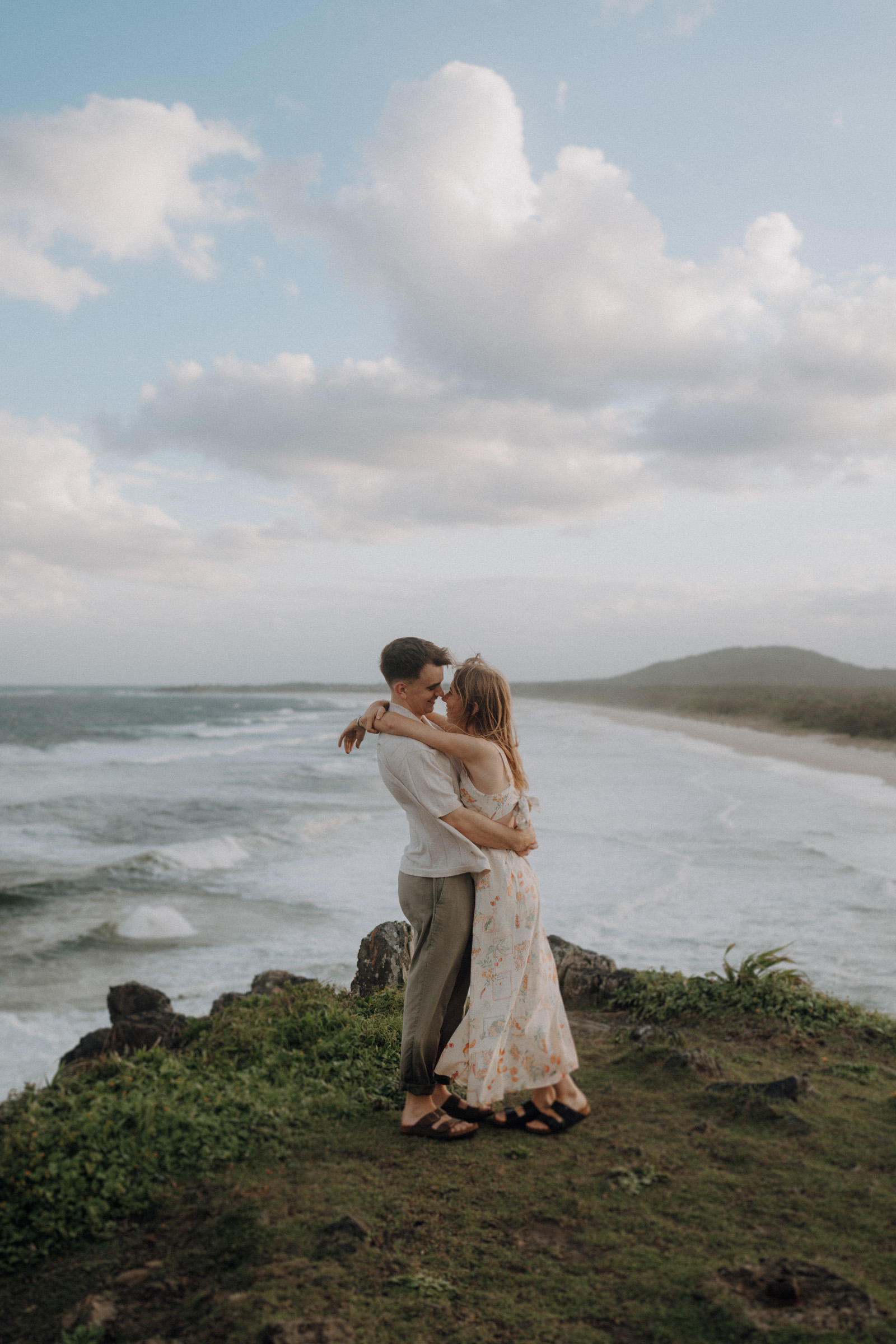 A couple embraces on a grassy cliff overlooking the ocean, under a cloudy sky.