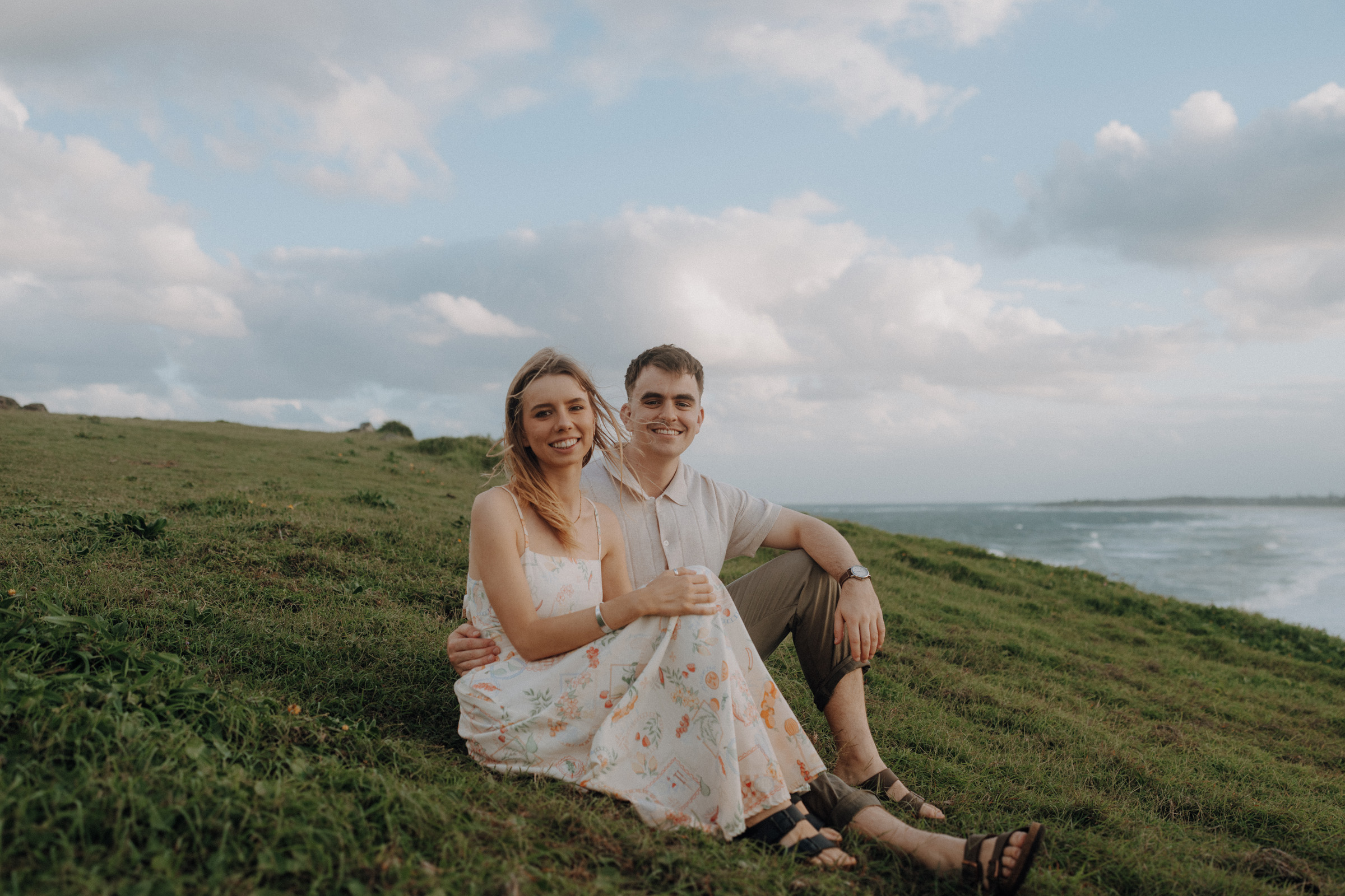 A couple sits on a grassy hillside near the ocean. The woman wears a floral dress, and the man is in a light shirt and pants. They are smiling with the sea and cloudy sky in the background.