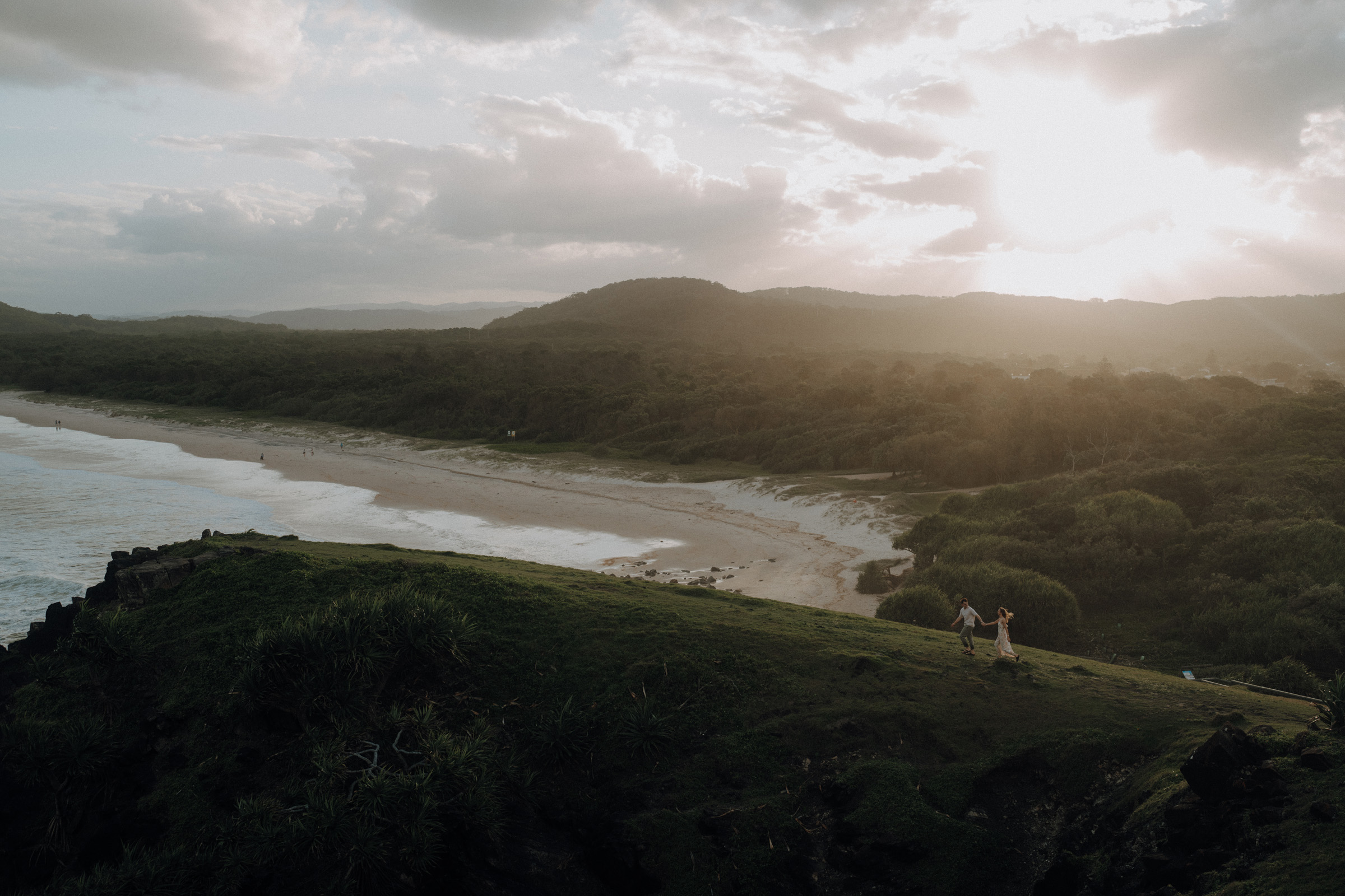 Two people walk along a grassy hill overlooking a sandy beach at sunset, with rolling hills and scattered clouds in the background.