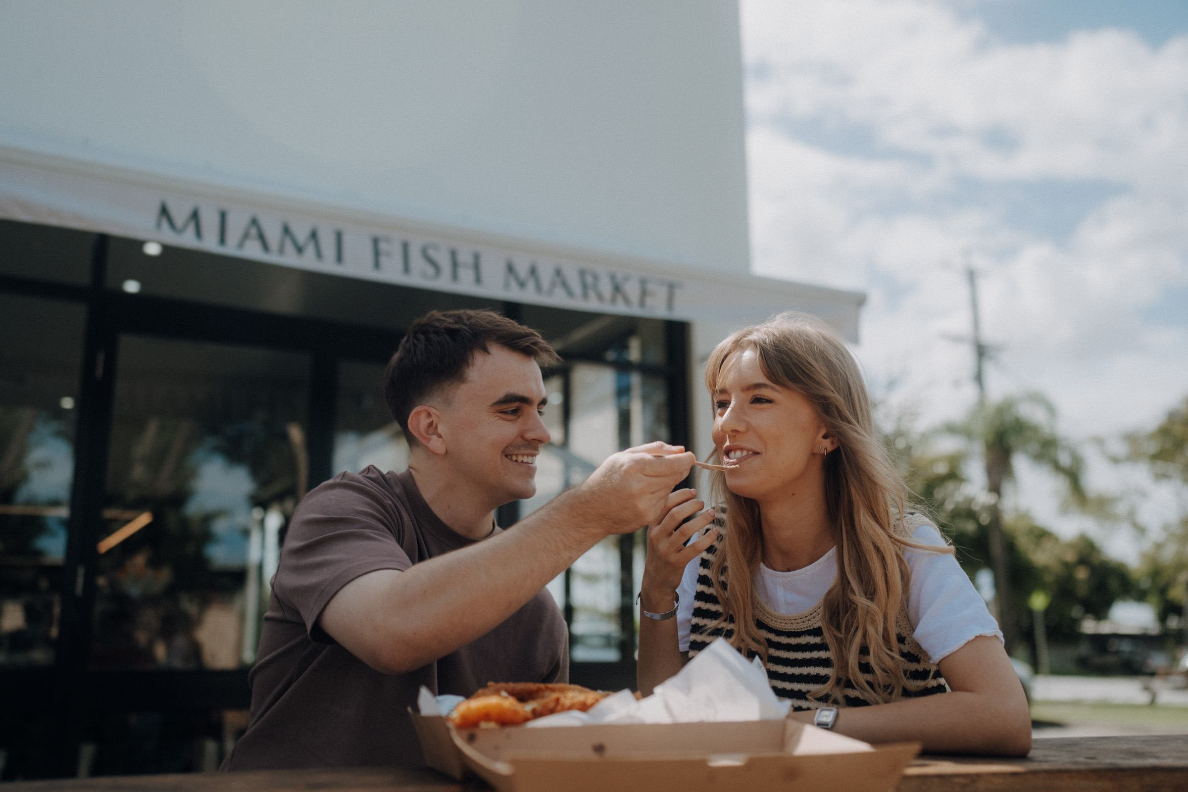 Two people sitting outside "Miami Fish Market." One person is feeding the other with a fork from a food container, both appear to be enjoying the moment.