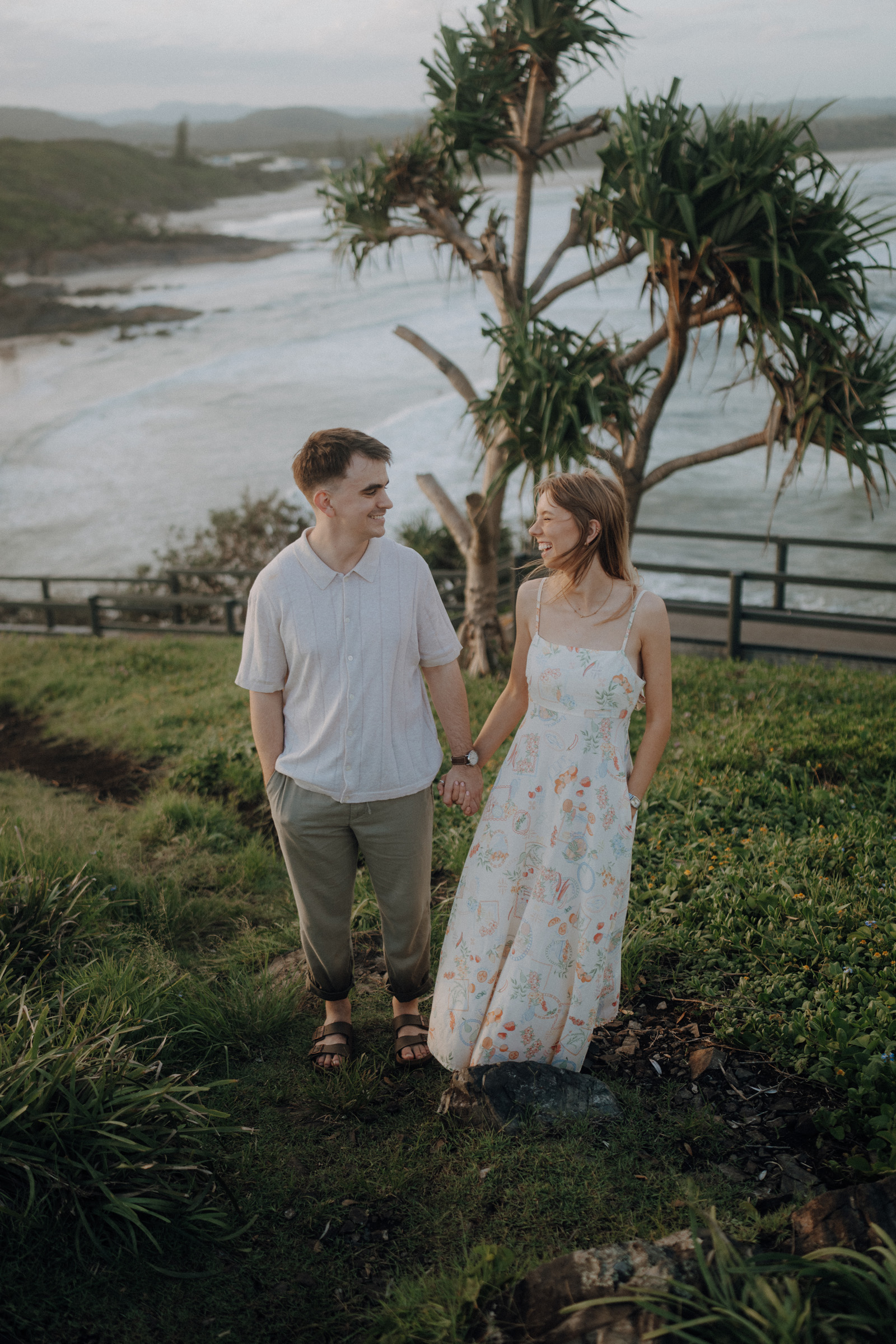 A man and a woman hold hands and walk along a grassy path by the seaside, with a tree and the ocean in the background.