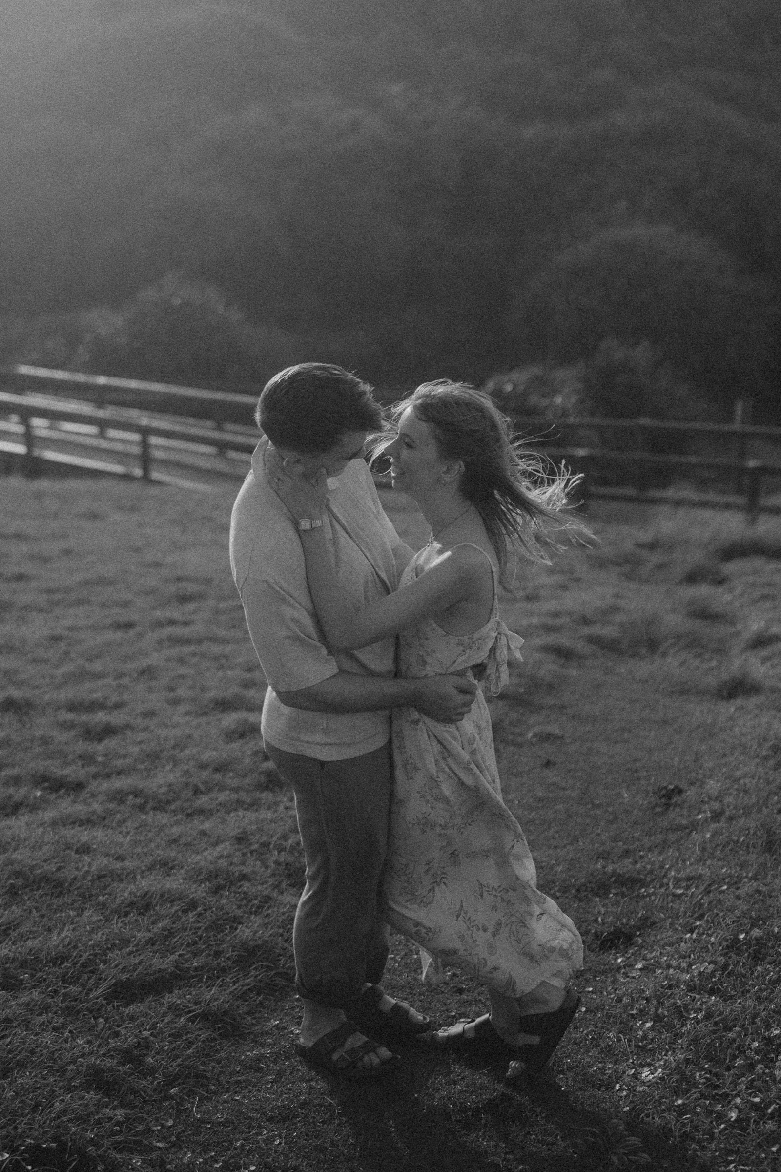 A couple embraces and smiles in a grassy field with a wooden fence and hills in the background. The image is in black and white.