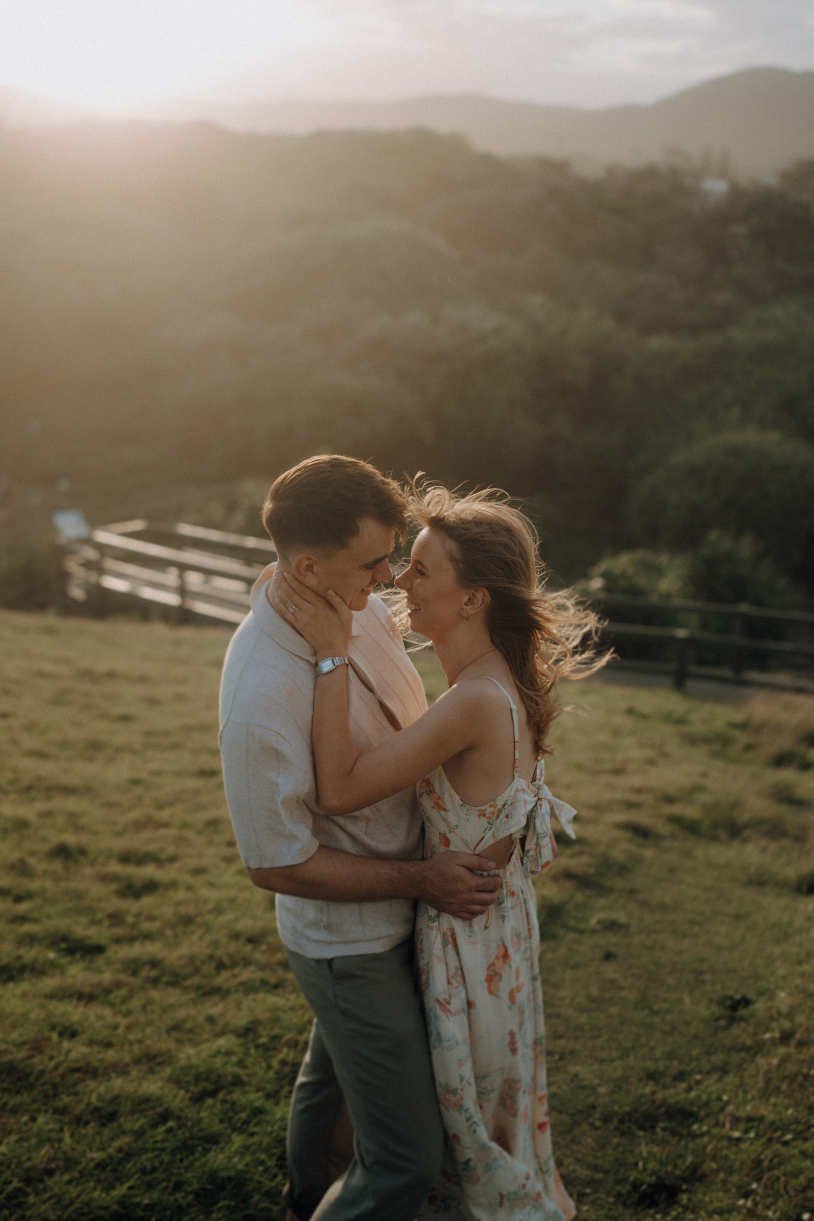 A couple embraces on a grassy hill during sunset, with lush greenery and hills in the background. The woman wears a floral dress and the man wears a light shirt.