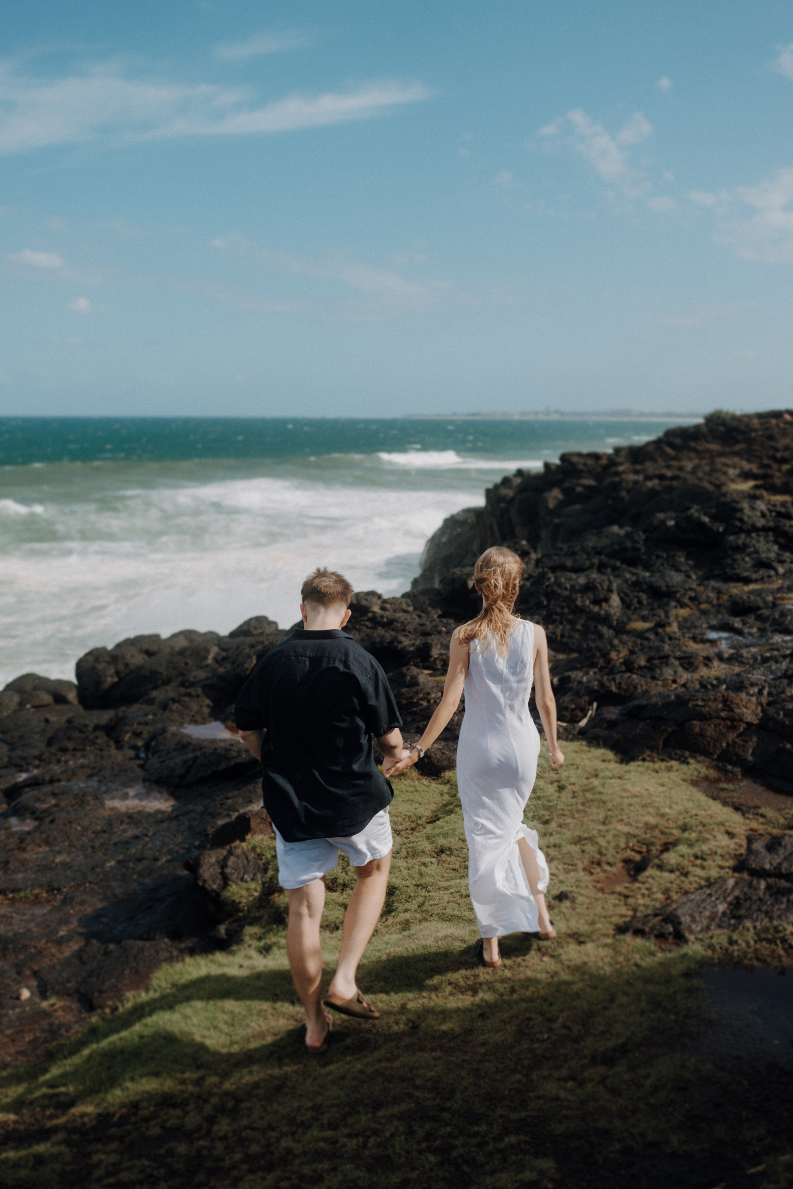 A couple, with a man in a black shirt and woman in a white dress, walks hand in hand on a grassy cliff overlooking the ocean, with rocks and waves in the background.