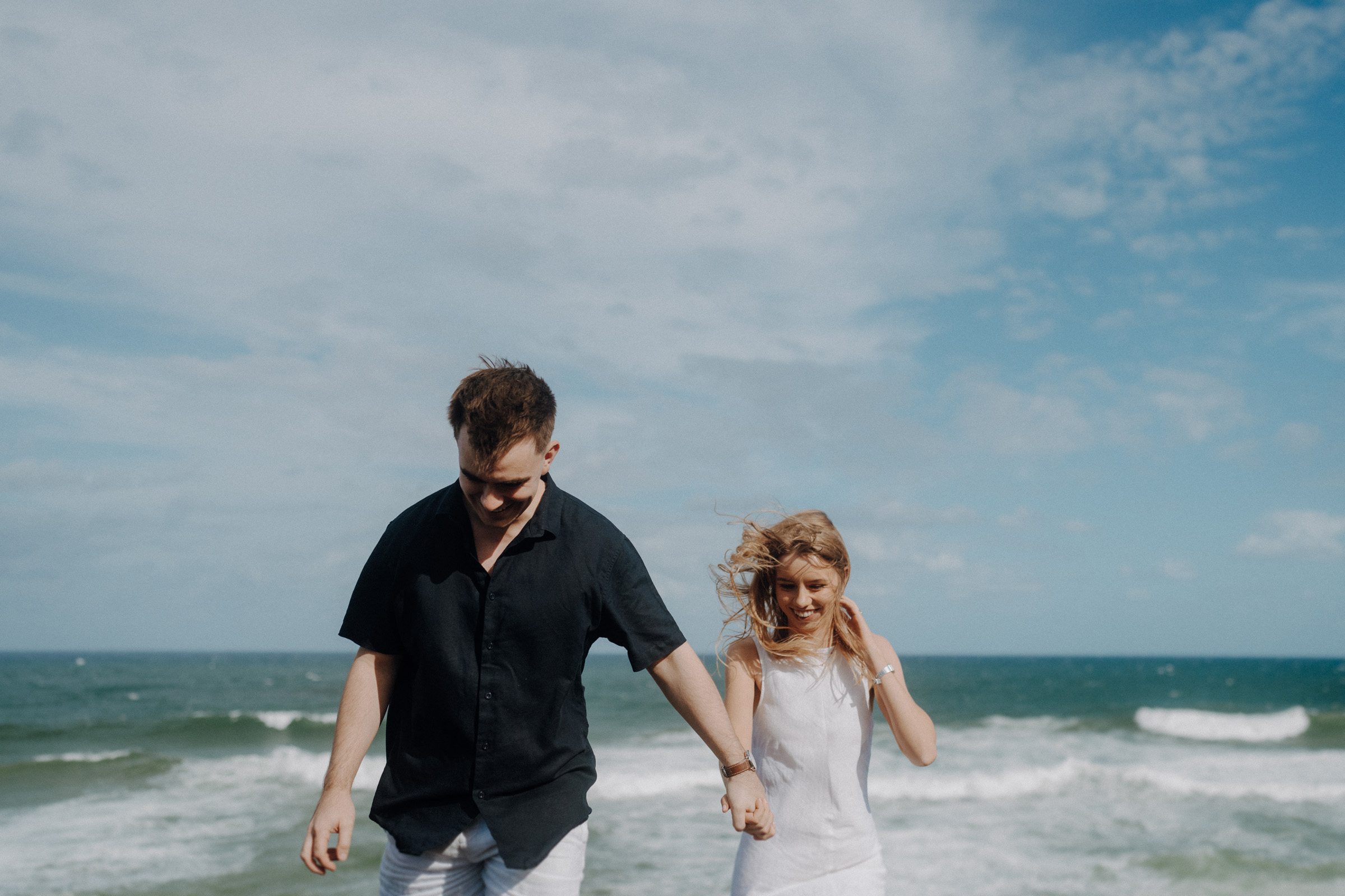 A man and woman hold hands while walking on a beach. The sky is partly cloudy, and waves are visible in the background.