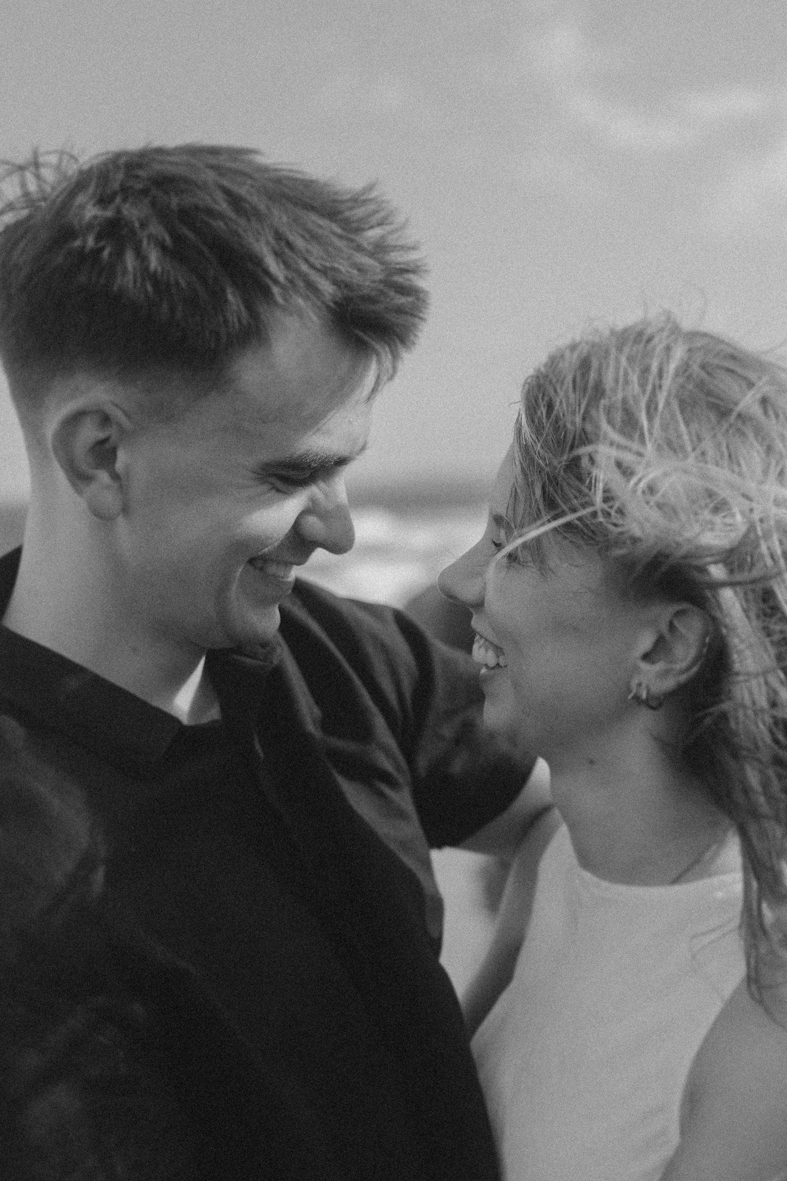 A black and white photo of a smiling couple standing close together, looking into each other's eyes. The man wears a dark shirt, and the woman wears a light top. It's a windy day.