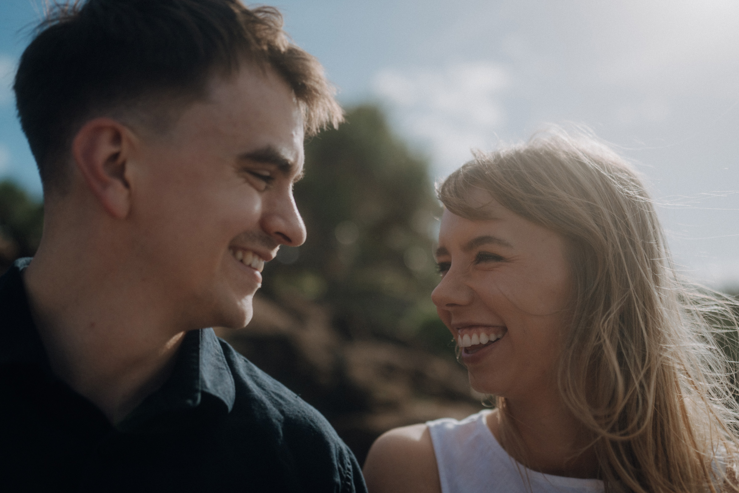 A man and a woman are smiling at each other outdoors with a bright sky and greenery in the background.