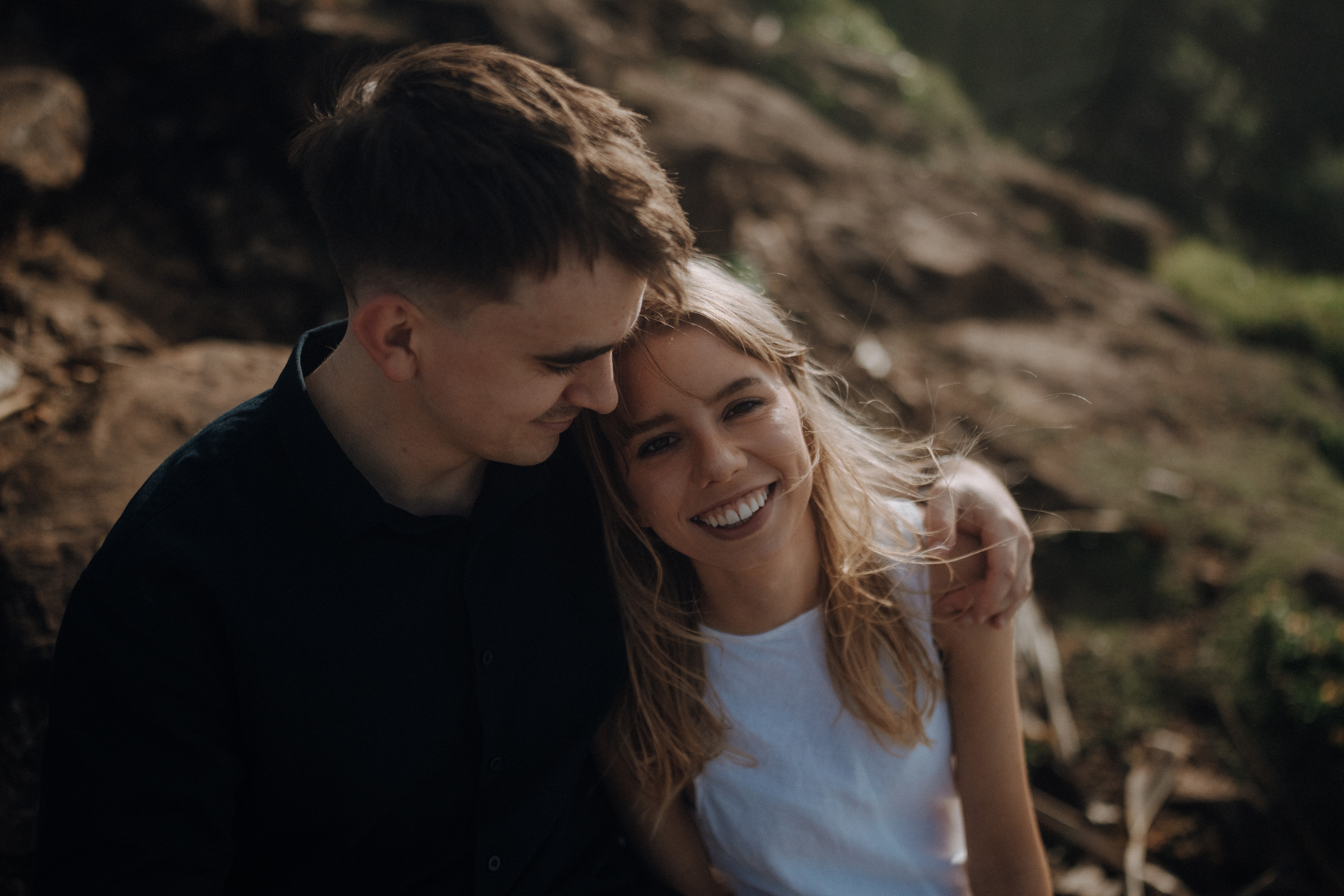 A couple sits closely on rocky ground. The man has his arm around the woman, who is smiling brightly. They are outdoors, surrounded by nature.