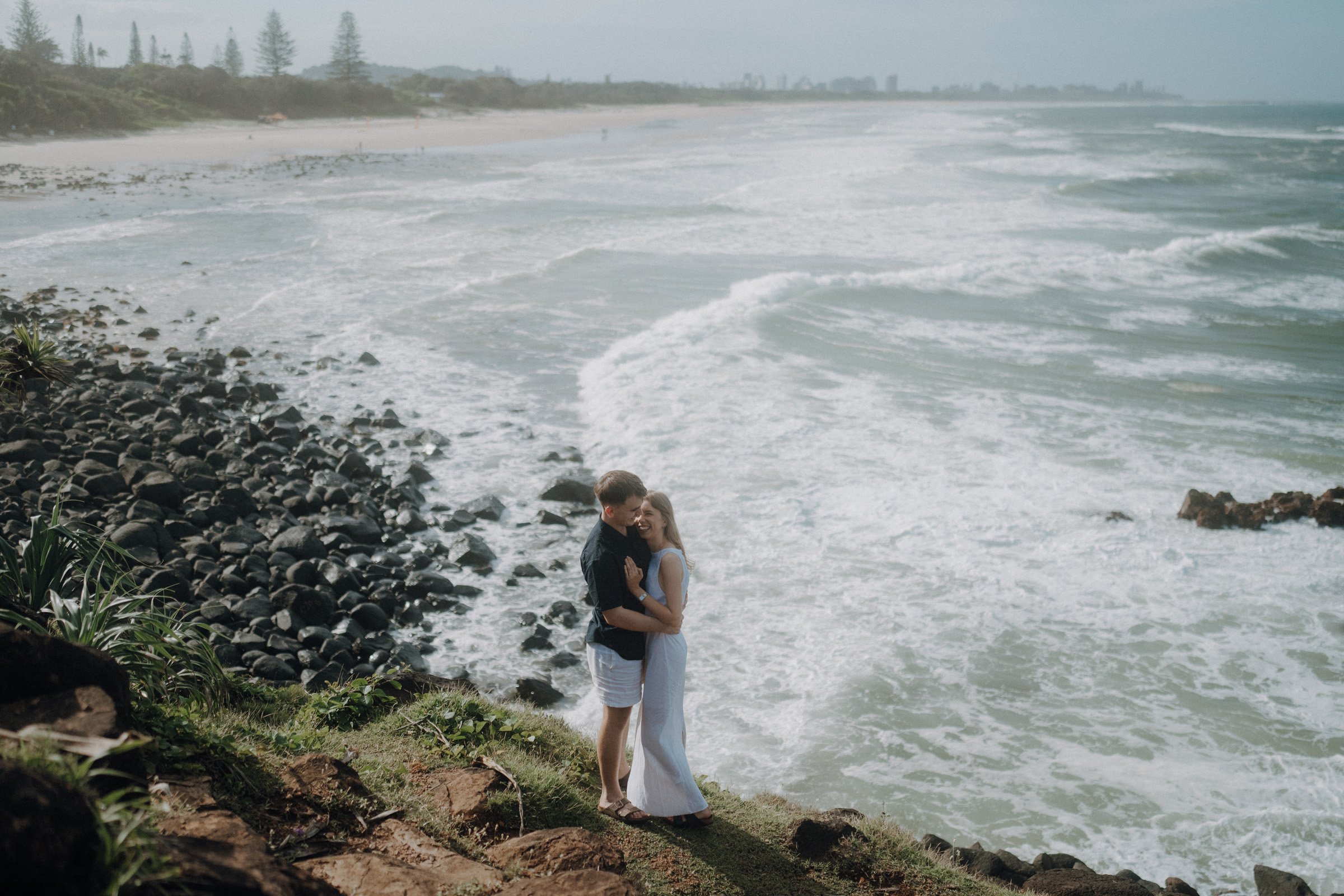 A couple embraces on a grassy cliff overlooking a rocky shoreline and waves, with a distant view of the beach and trees under a cloudy sky.