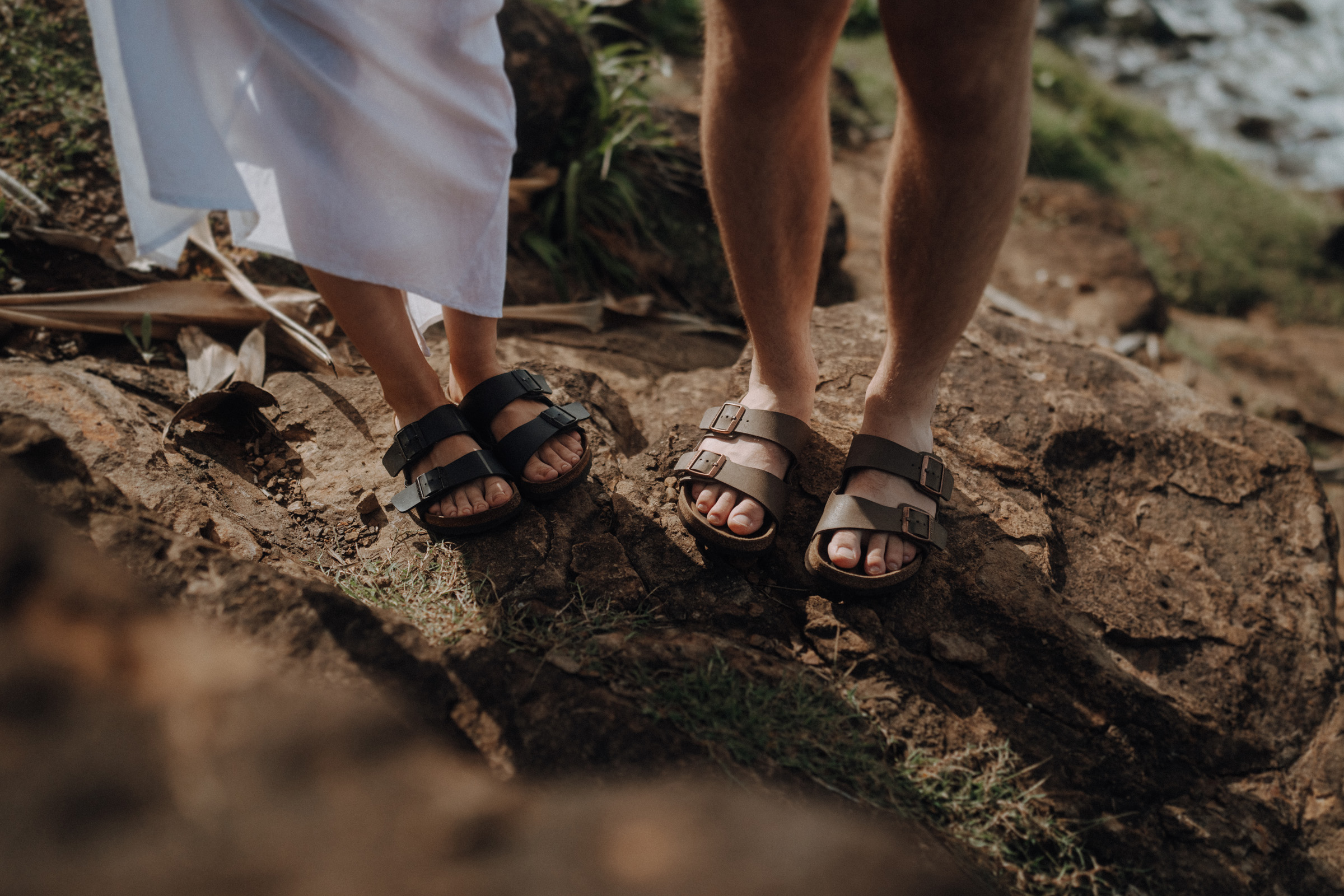 Two people stand on rocks near a grassy area, wearing sandals. One wears a white garment, and the other has shorts.