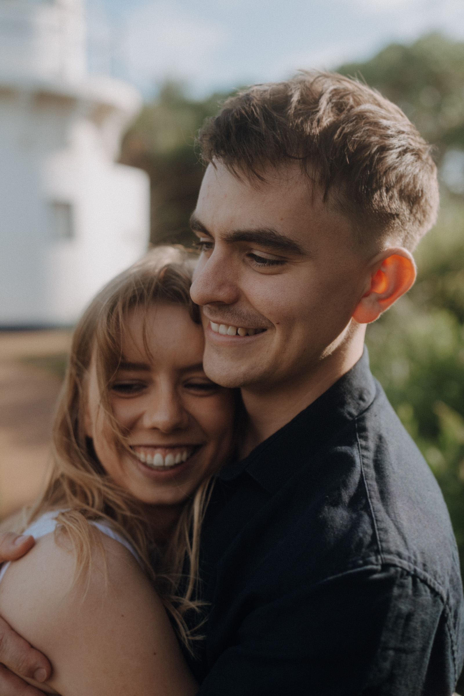 A smiling couple embraces outdoors with blurred greenery and a white building in the background.