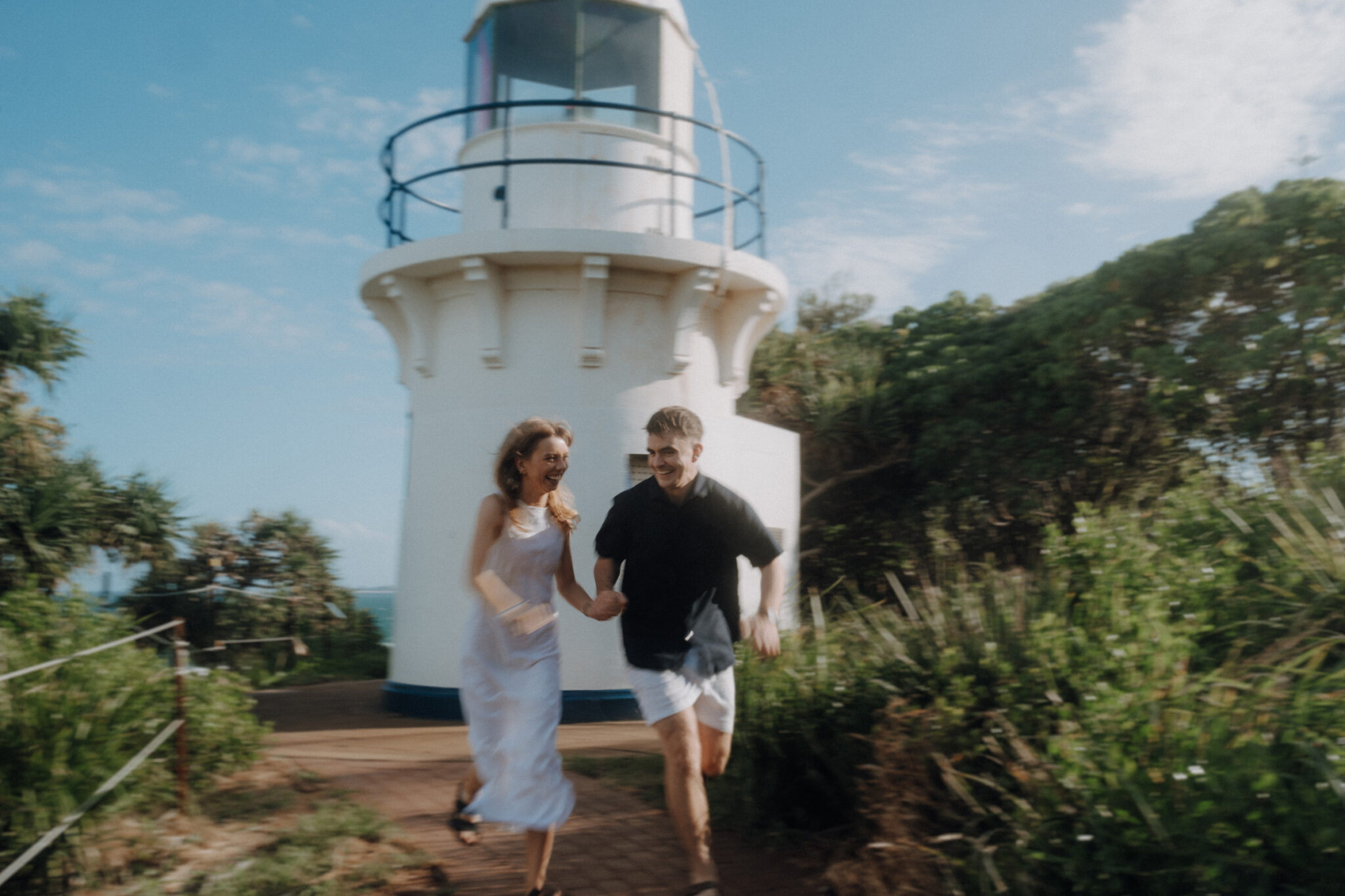 A man and woman joyfully run hand in hand near a white lighthouse surrounded by greenery.