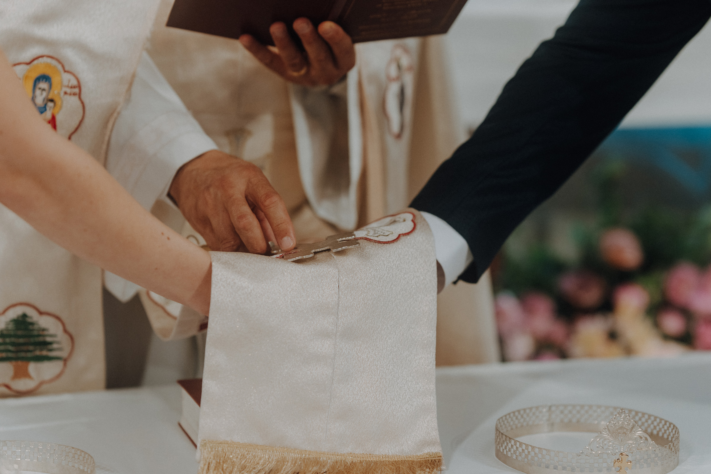 A bride and groom's hands are joined by a religious leader over a white cloth during a wedding ceremony. Two gold crowns are on the table.