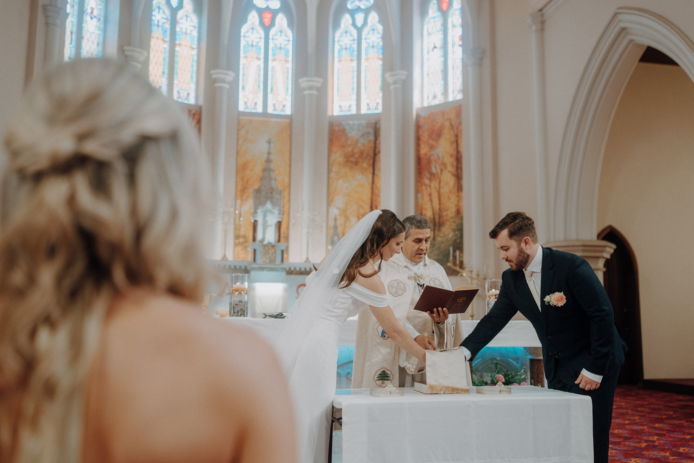 A bride and groom stand with a priest during a church wedding ceremony, surrounded by stained glass windows and arches.