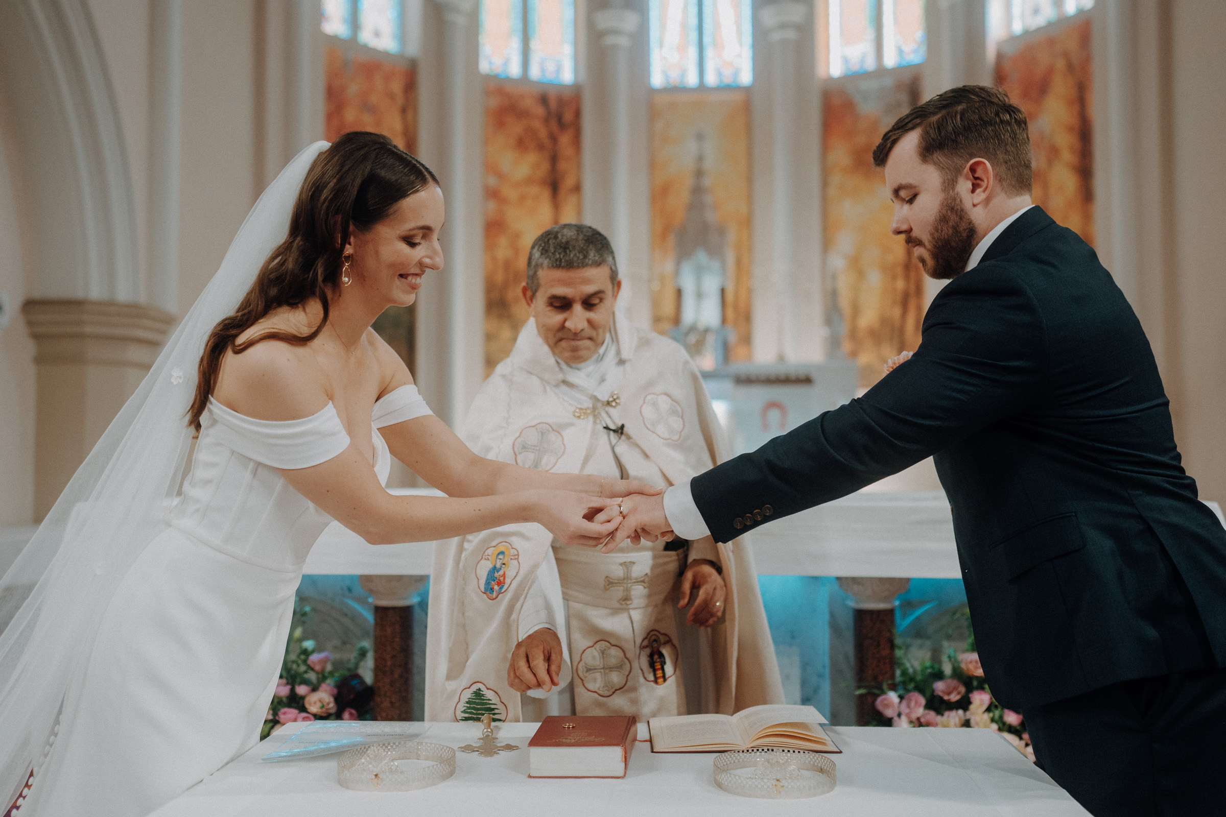 A bride and groom exchange rings in a church ceremony, with an officiant overseeing the event.