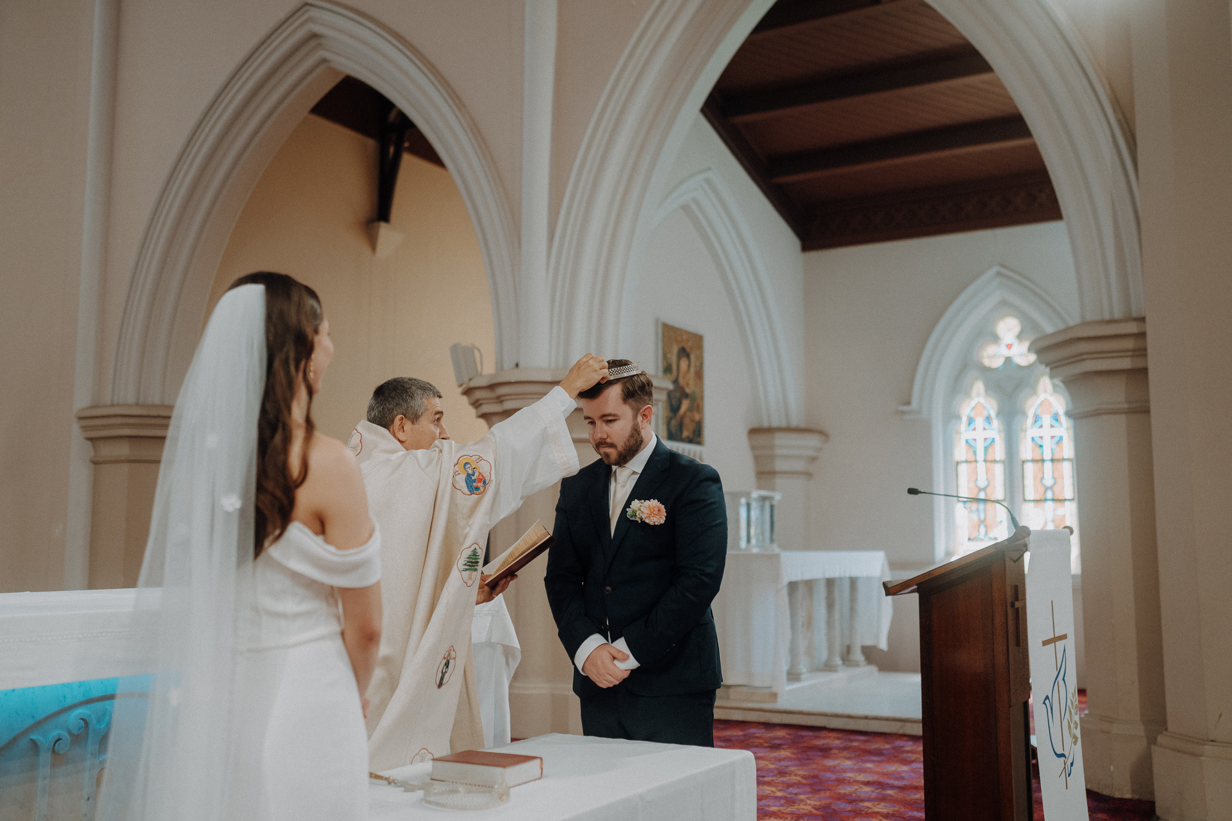 A priest blesses a groom during a wedding ceremony in a church, with the bride standing nearby.