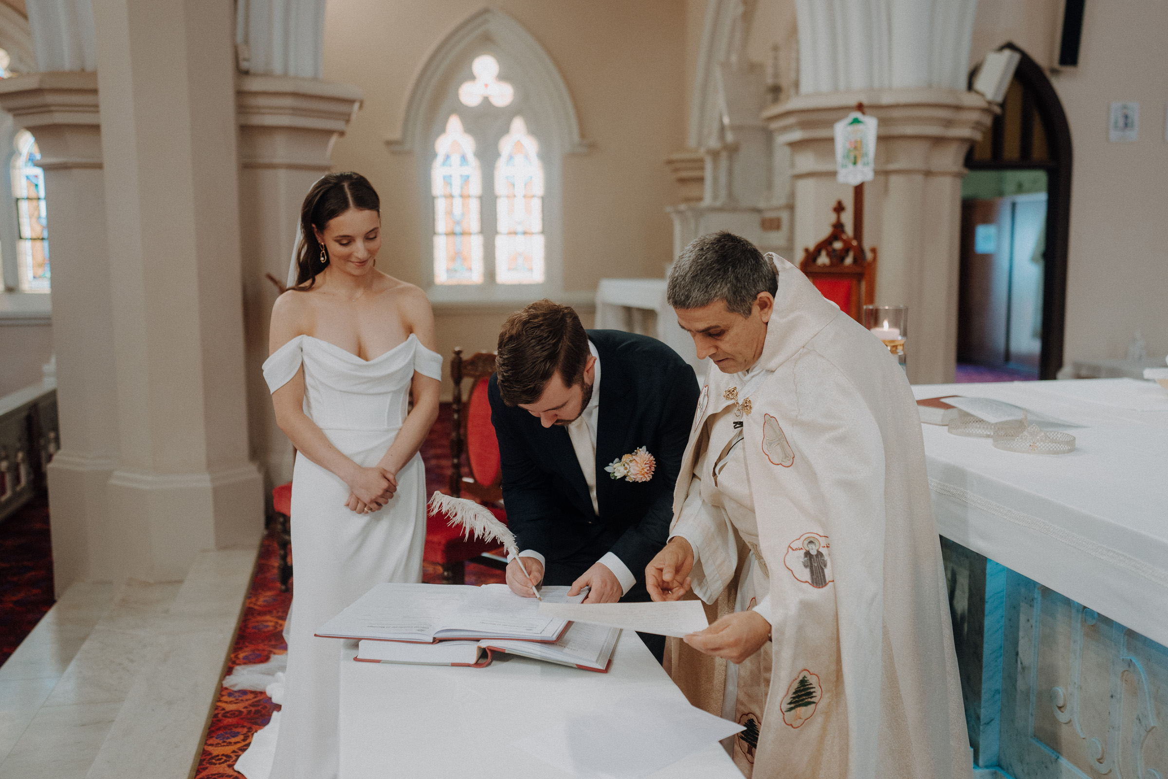 A groom signs a document on an altar during a wedding ceremony, while a bride stands nearby and a priest oversees the process inside a church.