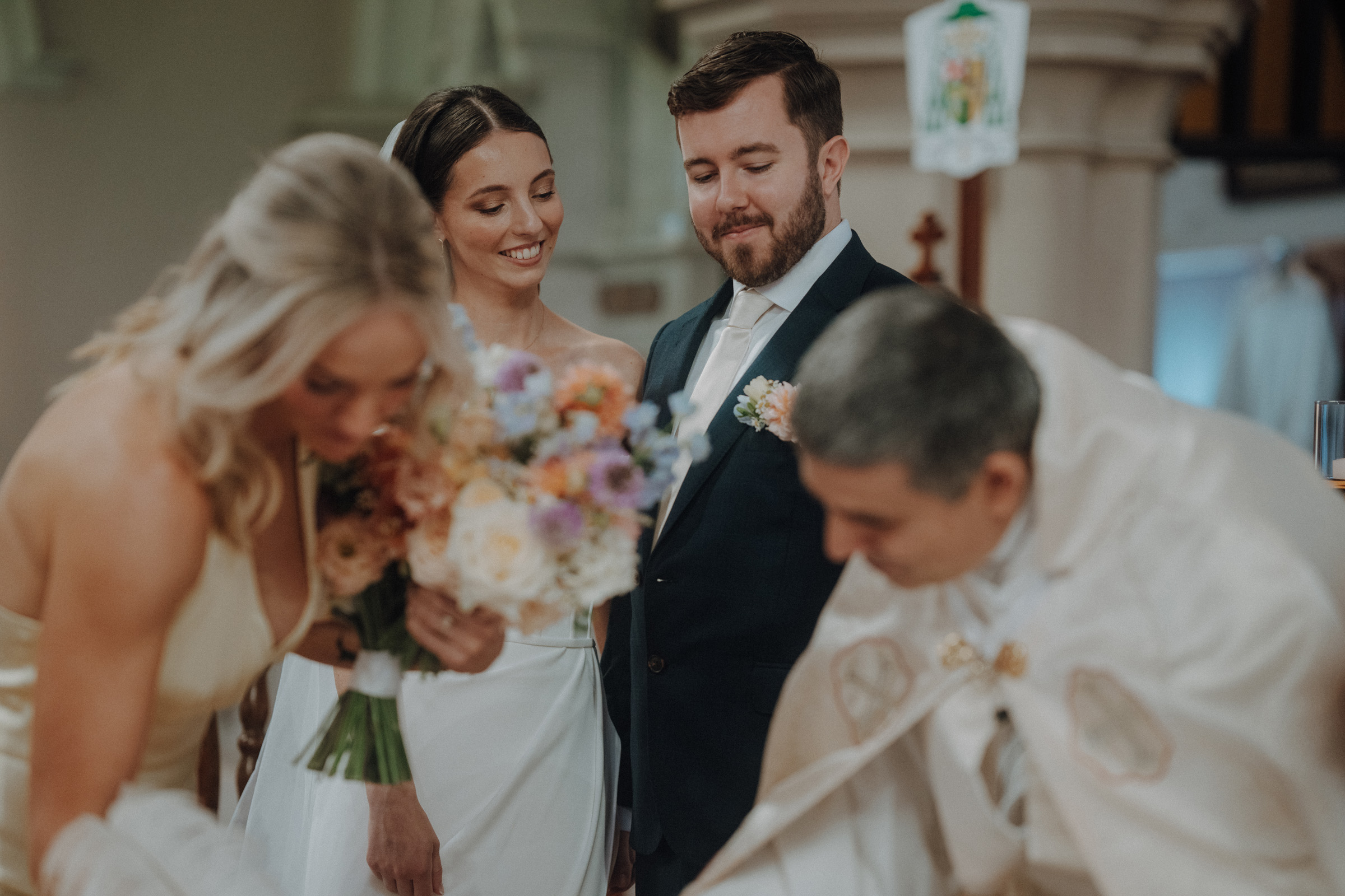 Bride and groom stand together during a wedding ceremony. A blonde woman holds flowers, and a clergyman in a white robe leans forward.