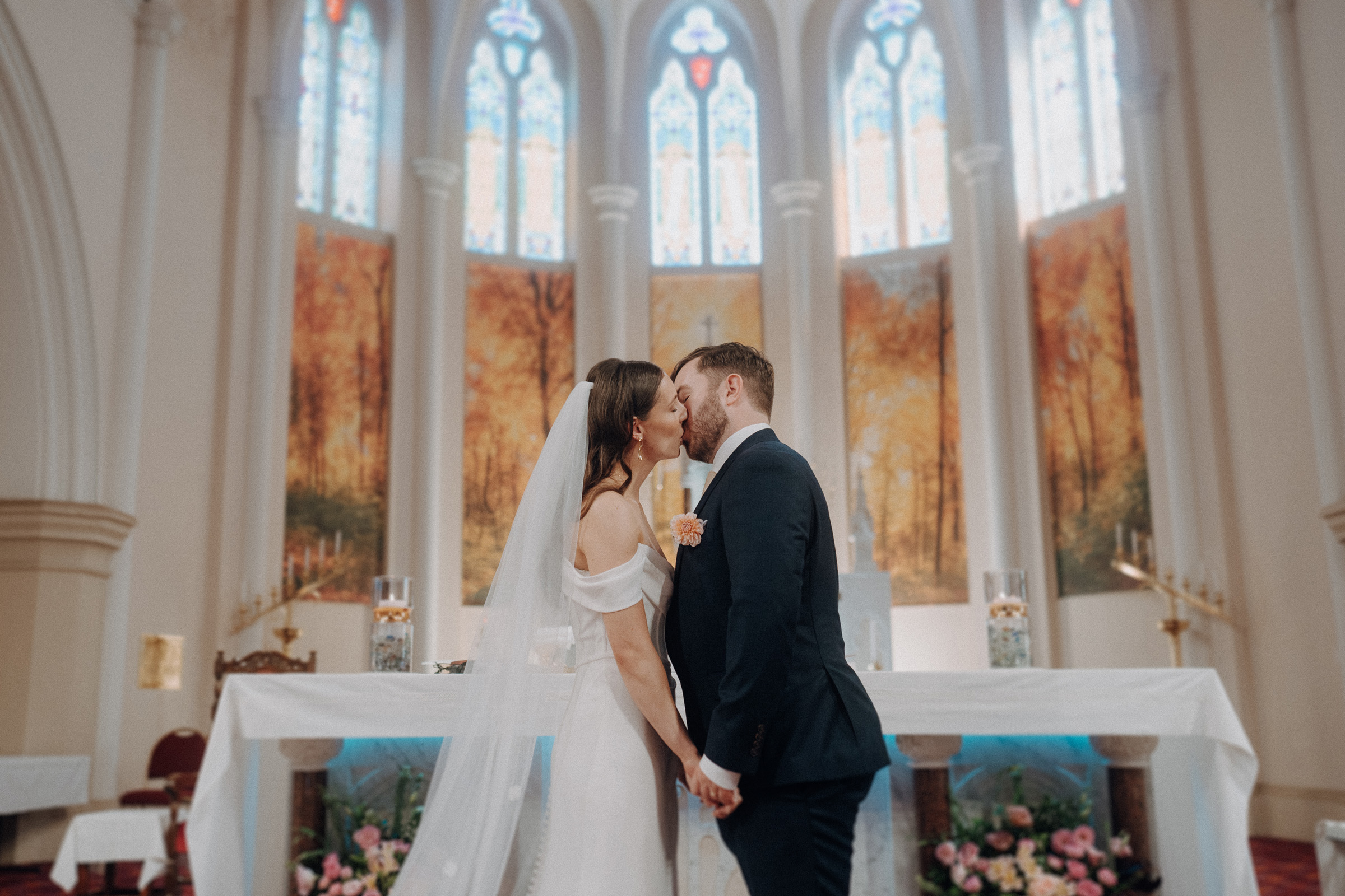 A bride and groom share a kiss in a church, standing before an altar decorated with pink flowers, with stained glass windows in the background.