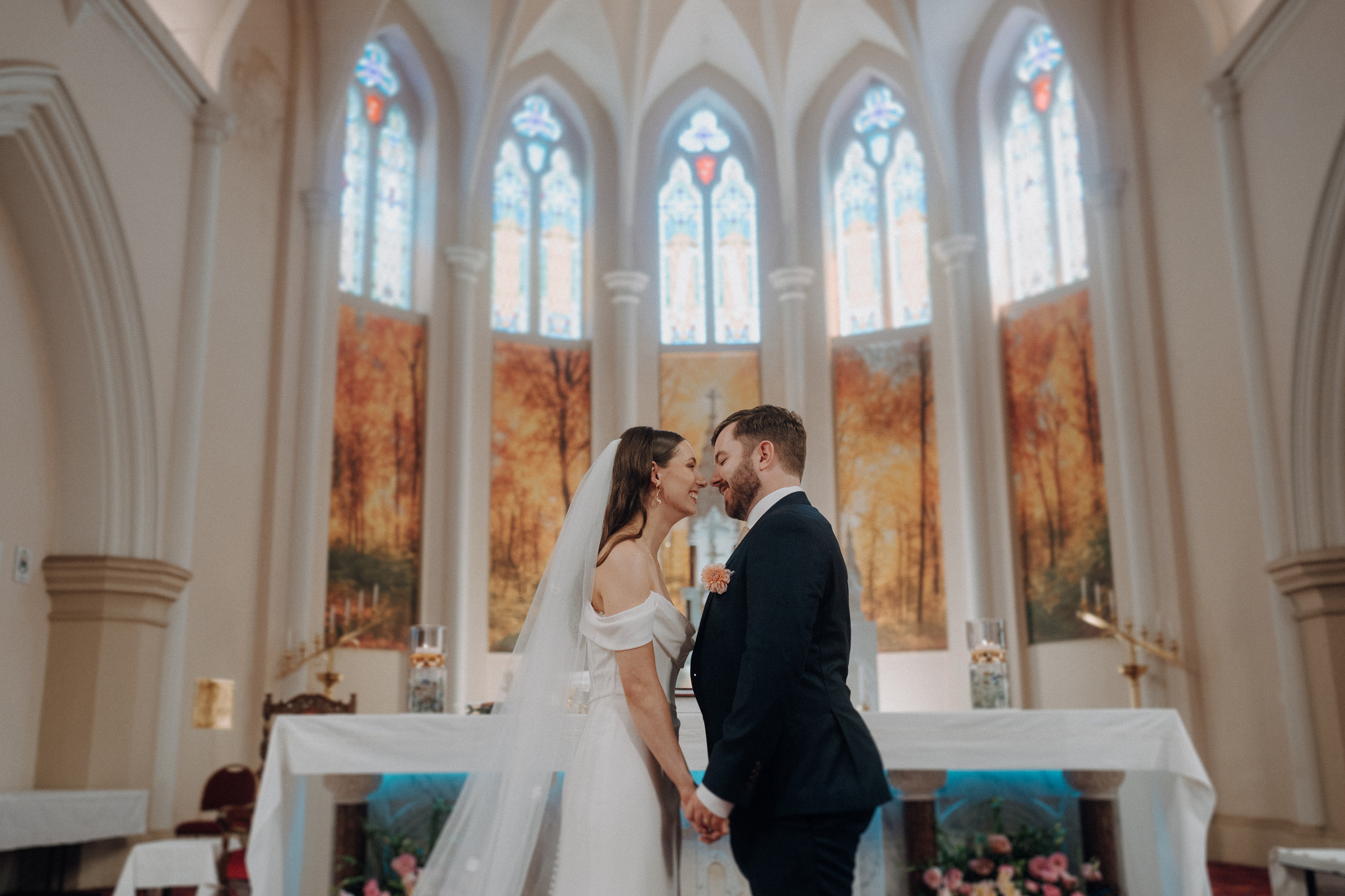 A bride and groom stand facing each other, holding hands inside a decorated church with stained glass windows.