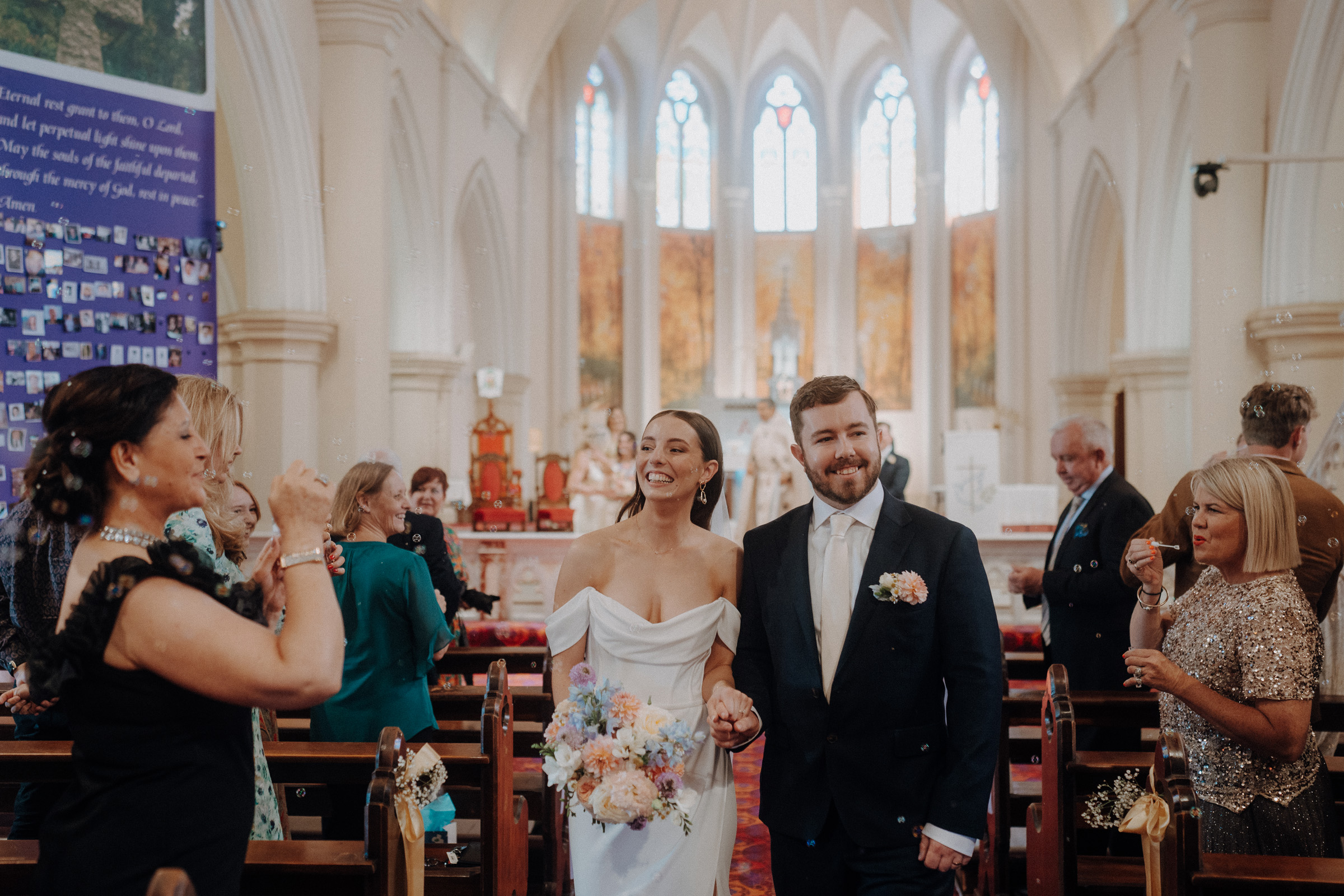 A bride and groom walk down the aisle of a church, smiling. Guests, some taking photos, are seated and standing around them. Stained glass windows and decorations are visible in the background.