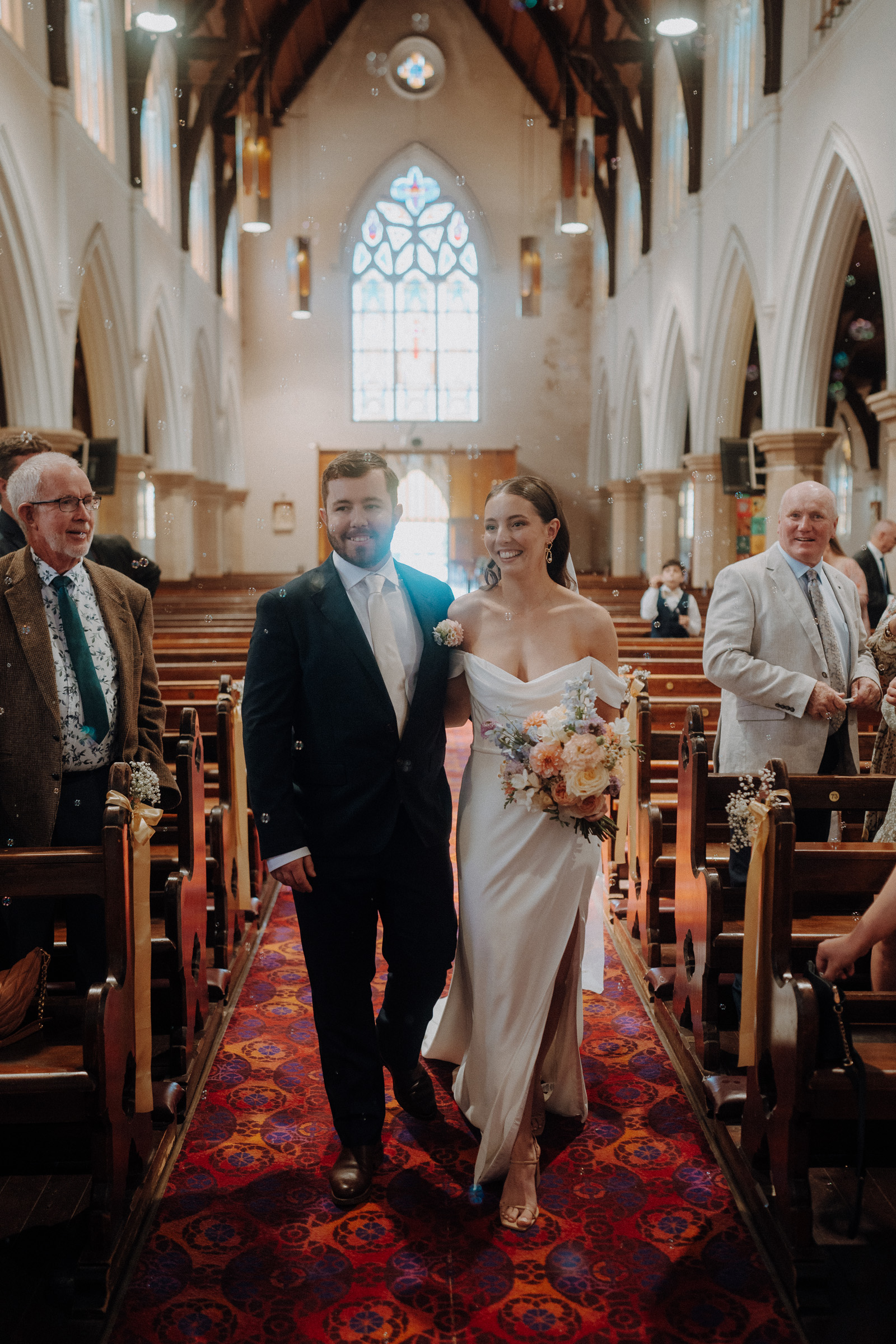 A bride and groom walk down the aisle of a church. The bride is holding a bouquet, and guests are seated on either side. Stained glass windows are visible in the background.