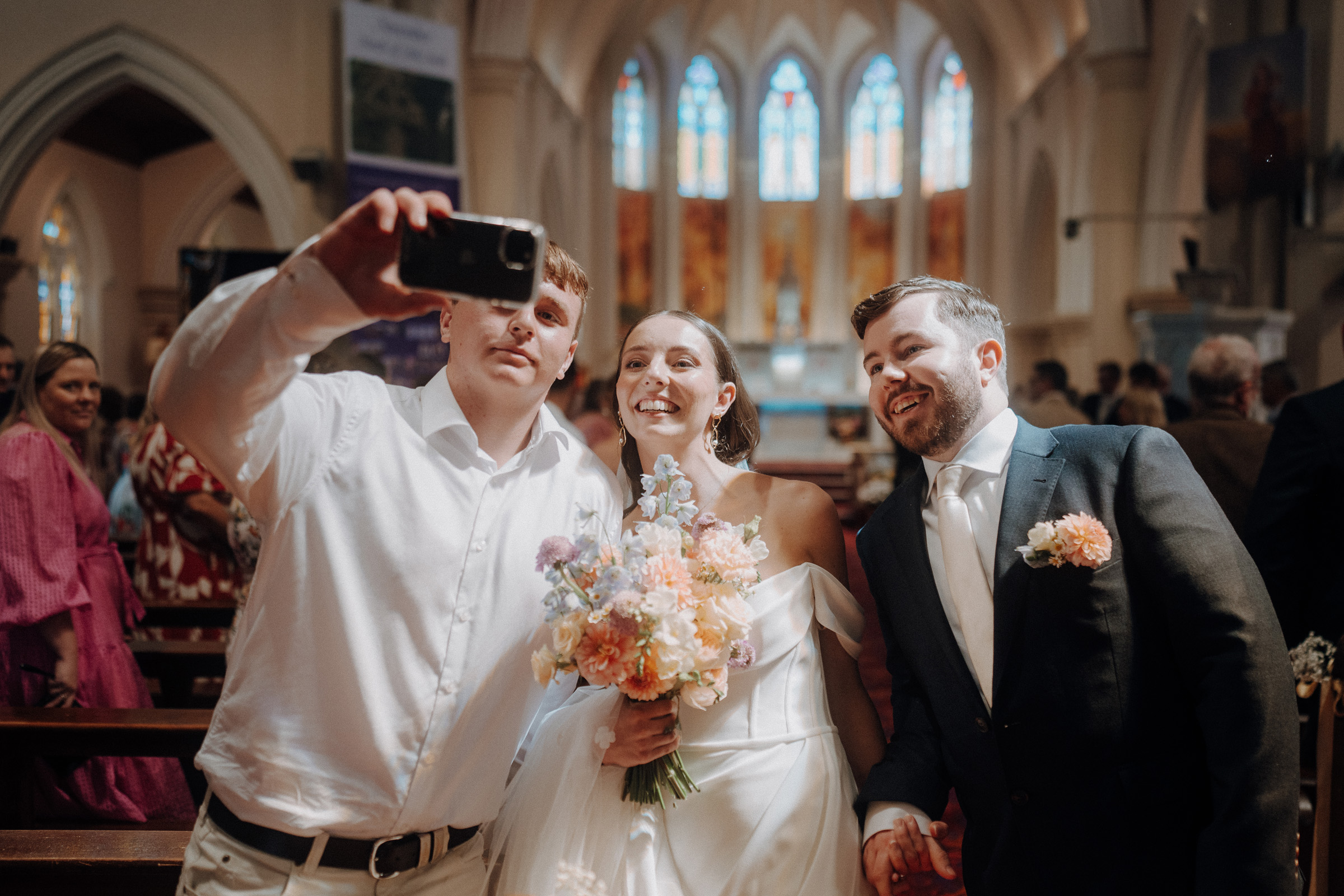 A bride and groom pose for a selfie with a guest in a church. The bride holds a bouquet of flowers, and colorful stained-glass windows are visible in the background.