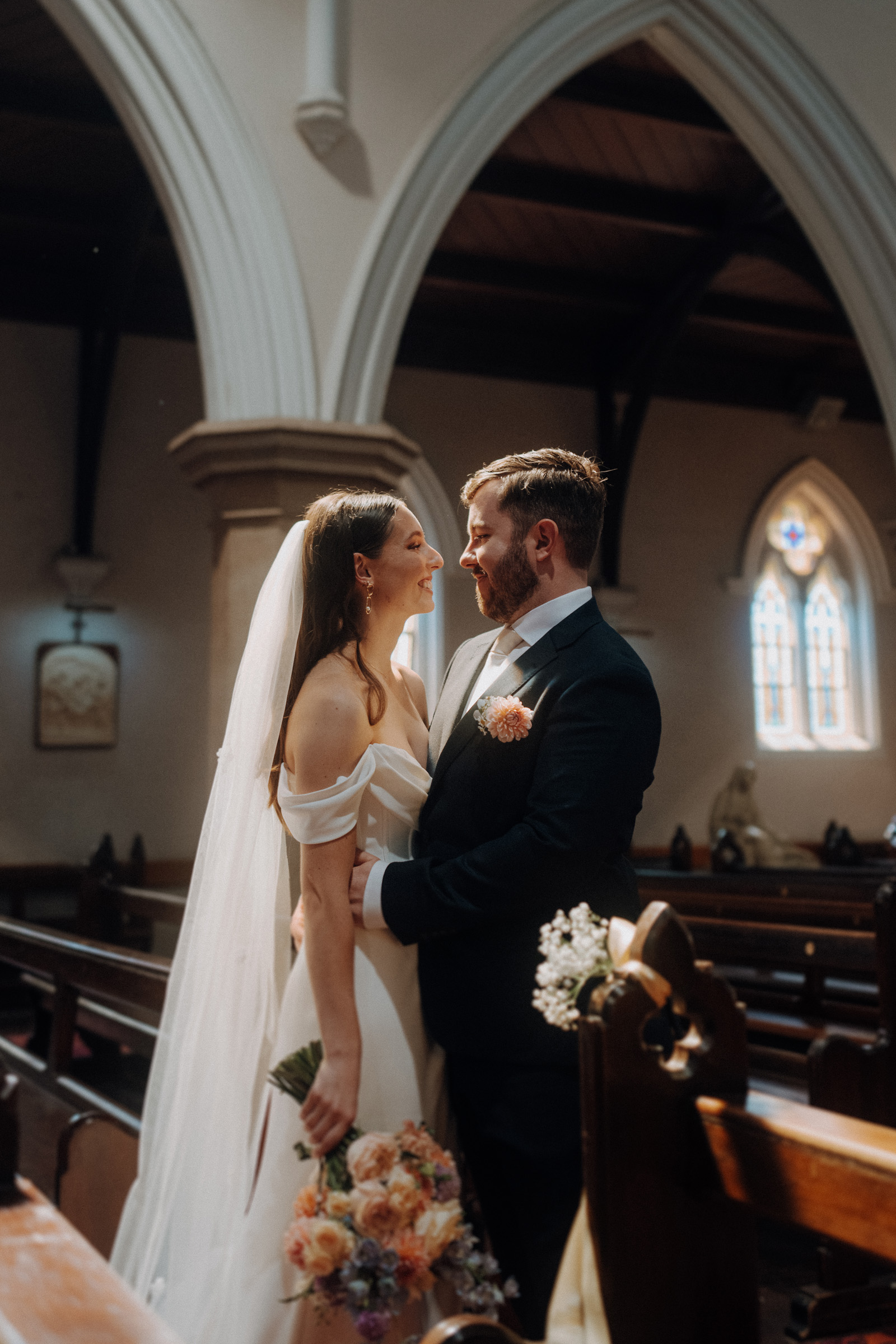 A bride and groom stand together inside a church, gazing at each other. The bride holds a bouquet of flowers, and light streams through stained glass windows.