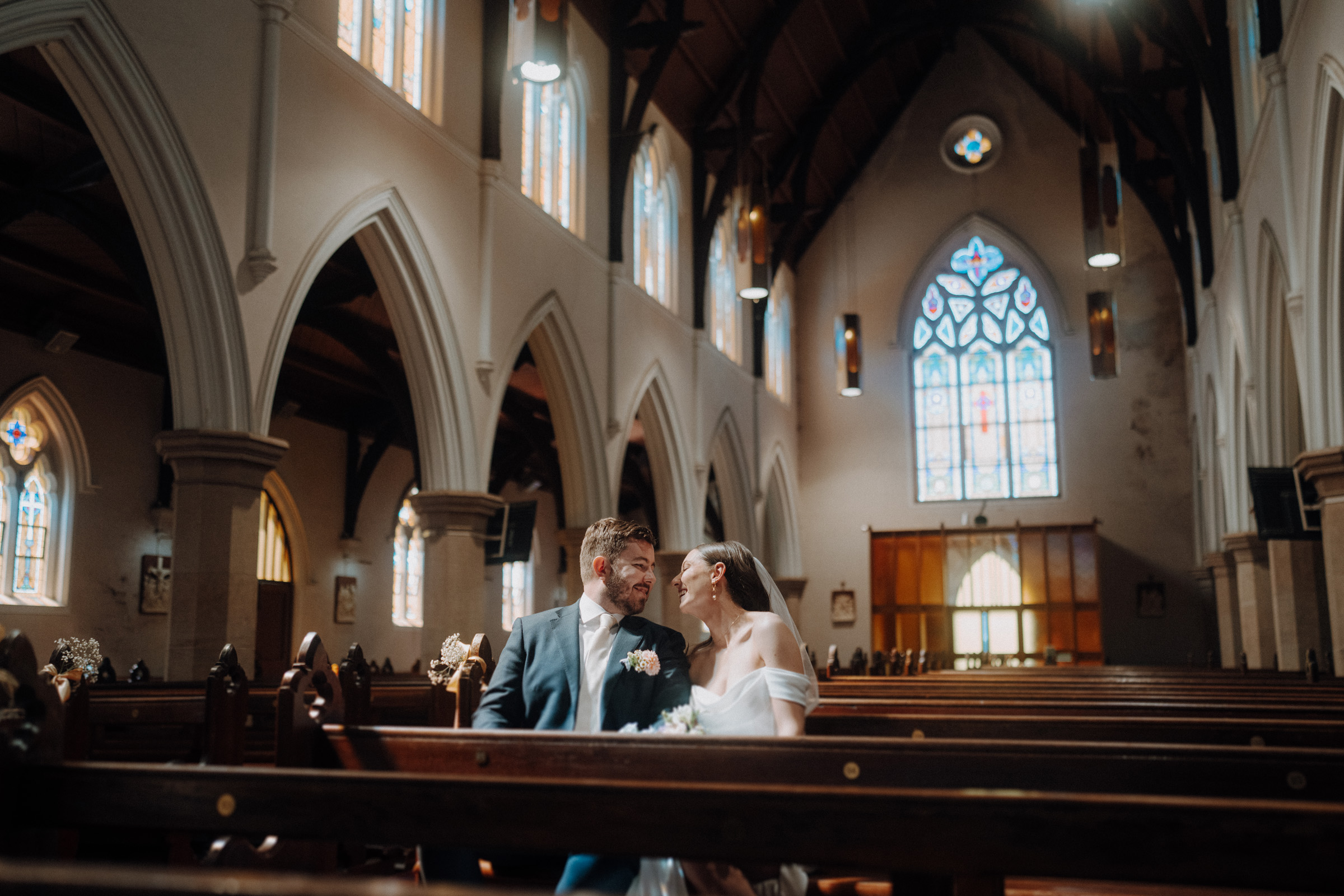 A bride and groom sit in a church pew, looking at each other with smiles. The church has high arched ceilings and stained glass windows.