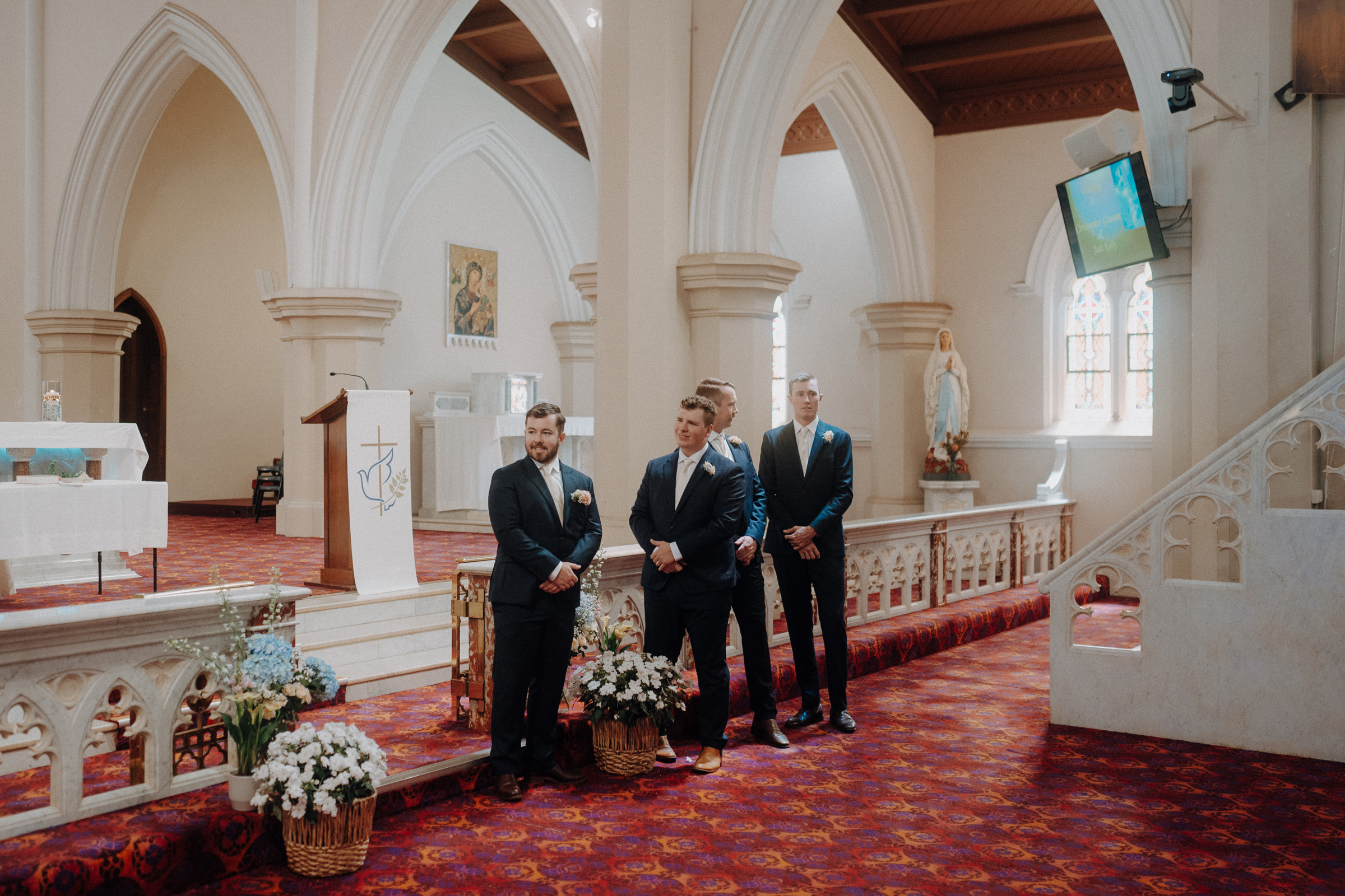 Three men in suits stand at the front of a church with arched architecture, near floral arrangements and a podium.