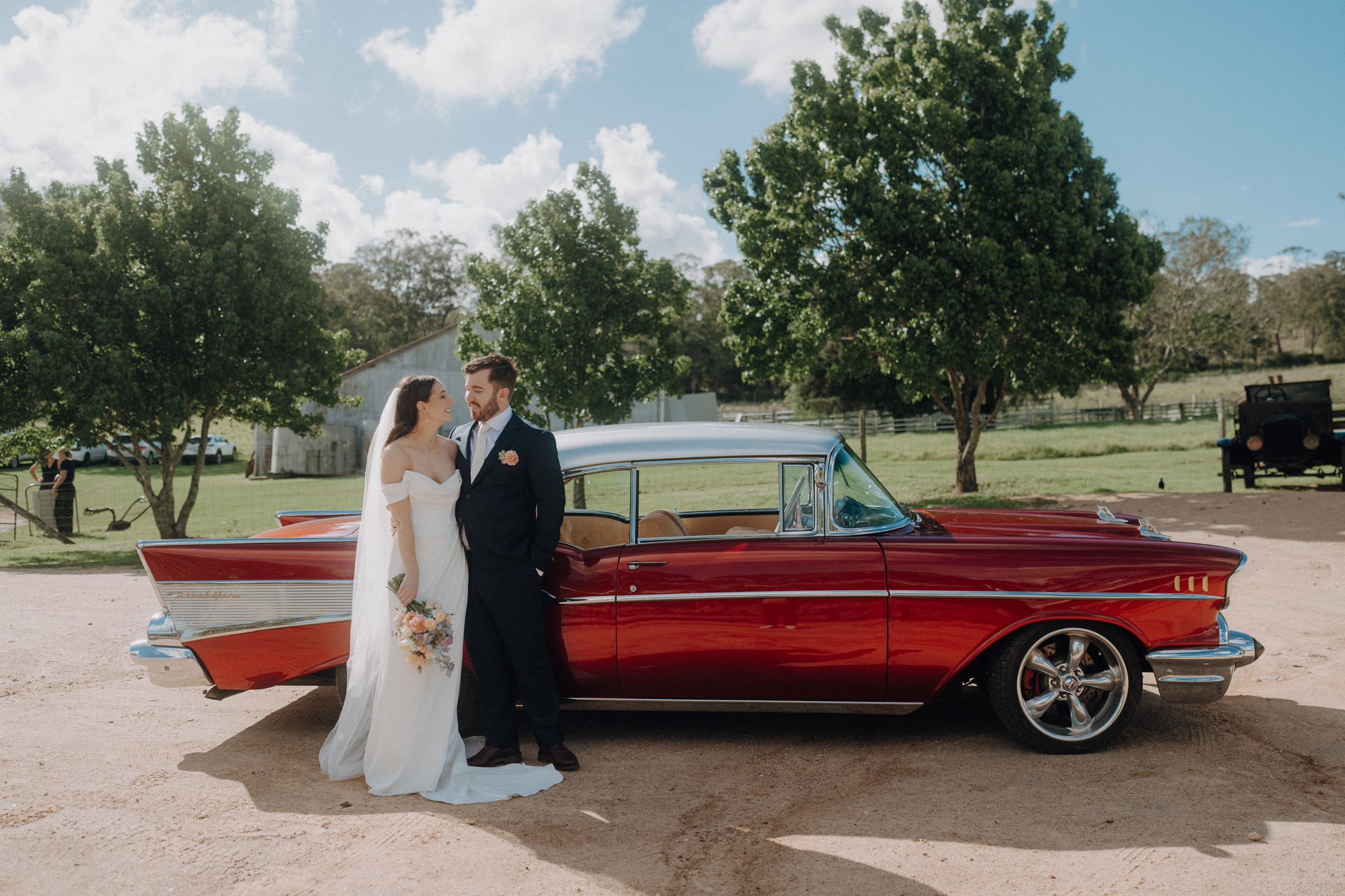 A bride and groom stand beside a vintage red car on a sunny day, with trees in the background.