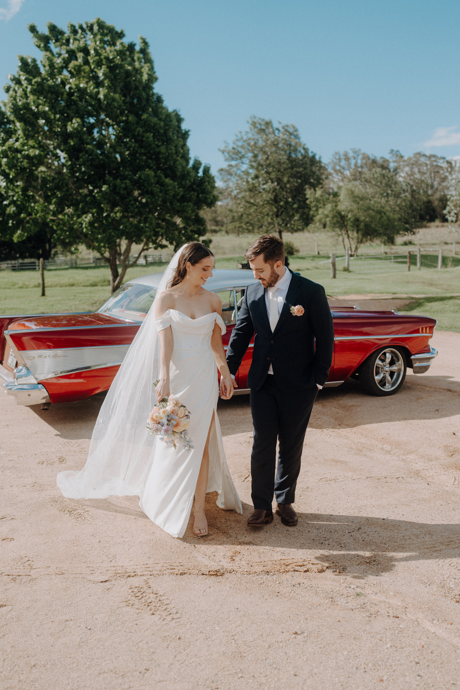 Bride and groom walking hand in hand on a sunny day, near a red vintage car, with trees and grass in the background.