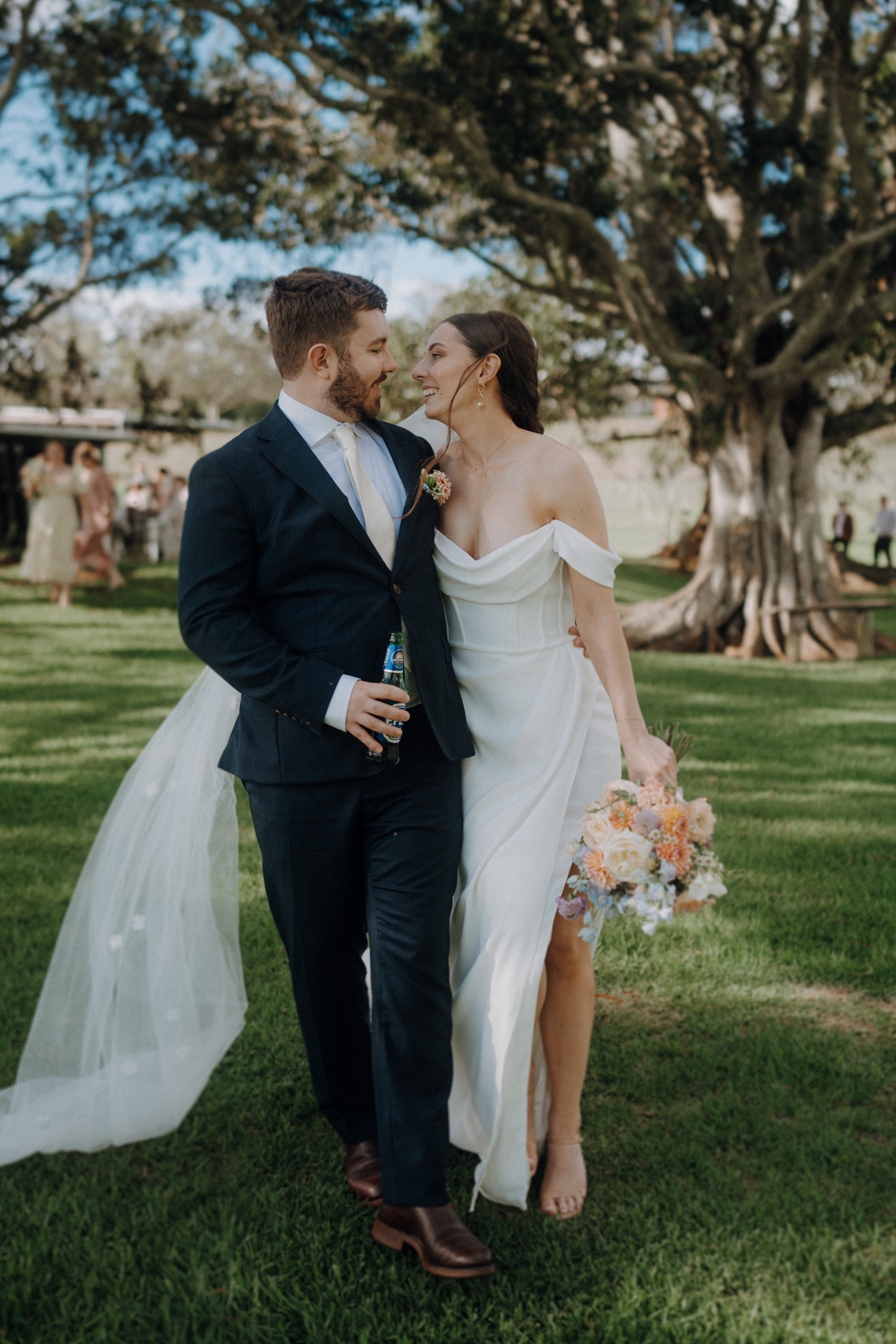 Bride and groom walk on grass, wearing formal attire. The groom holds a bottle, and the bride holds a bouquet. A large tree and people are in the background.