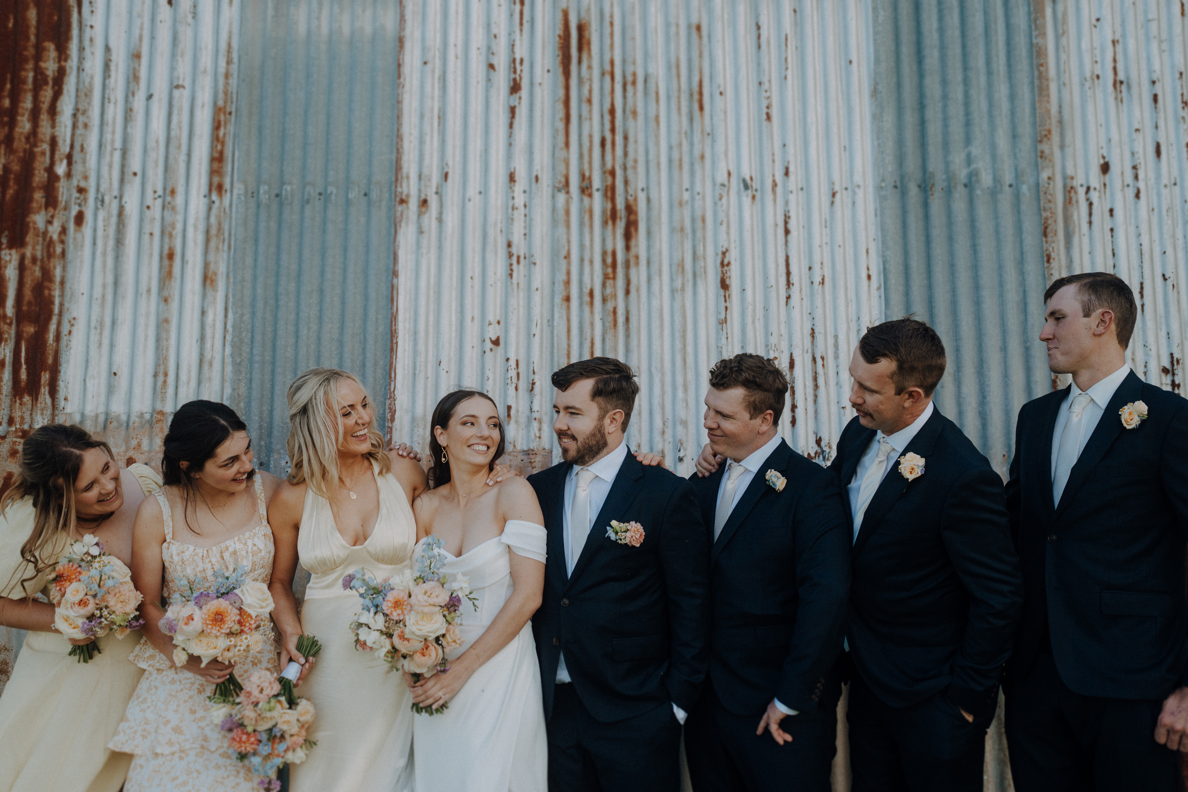 A wedding party poses in front of a rustic, corrugated metal wall. The bride and groom stand in the center, with bridesmaids holding bouquets to their left and groomsmen to their right.