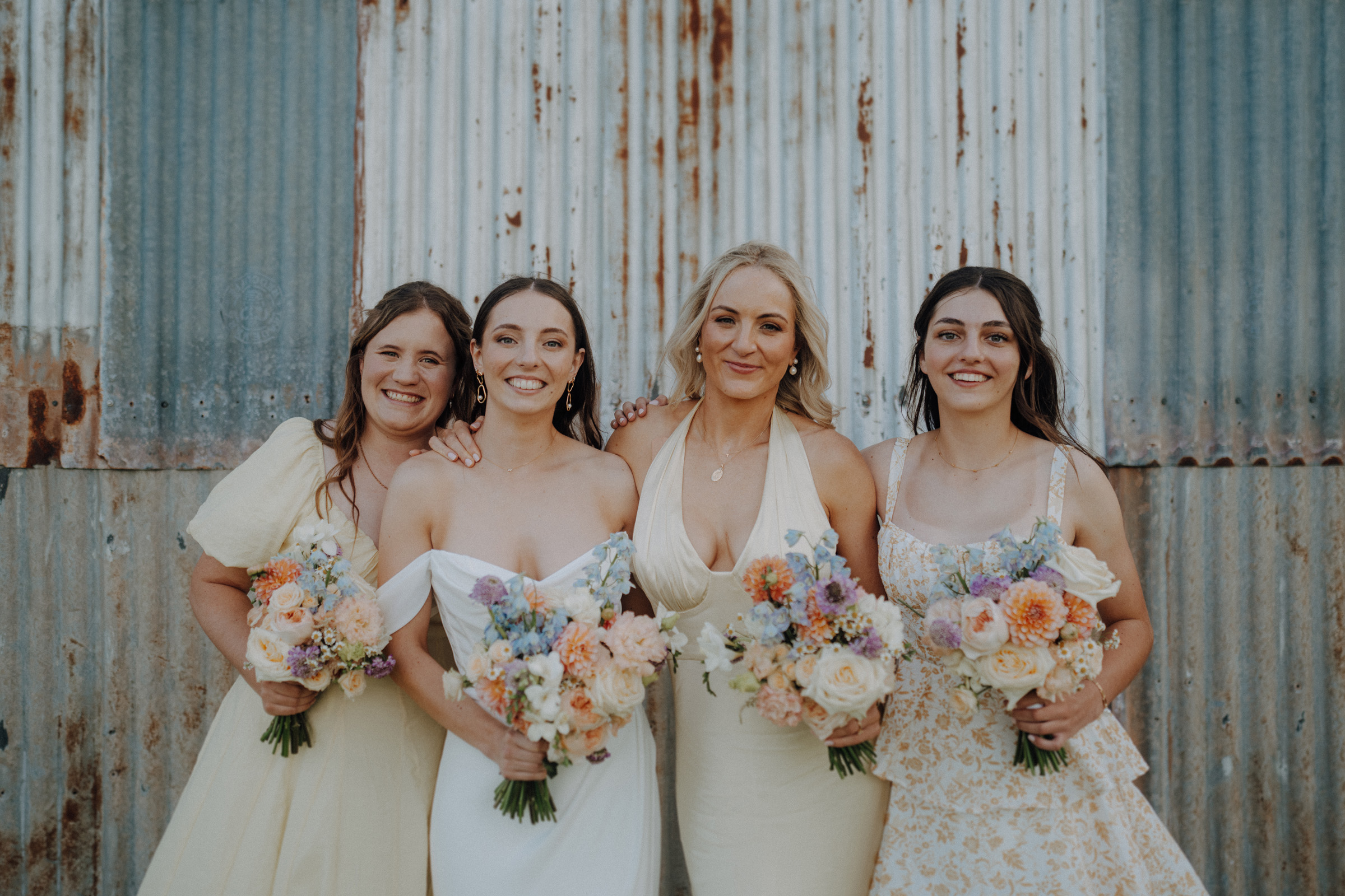 Four women in dresses stand smiling, holding bouquets, in front of a corrugated metal wall.