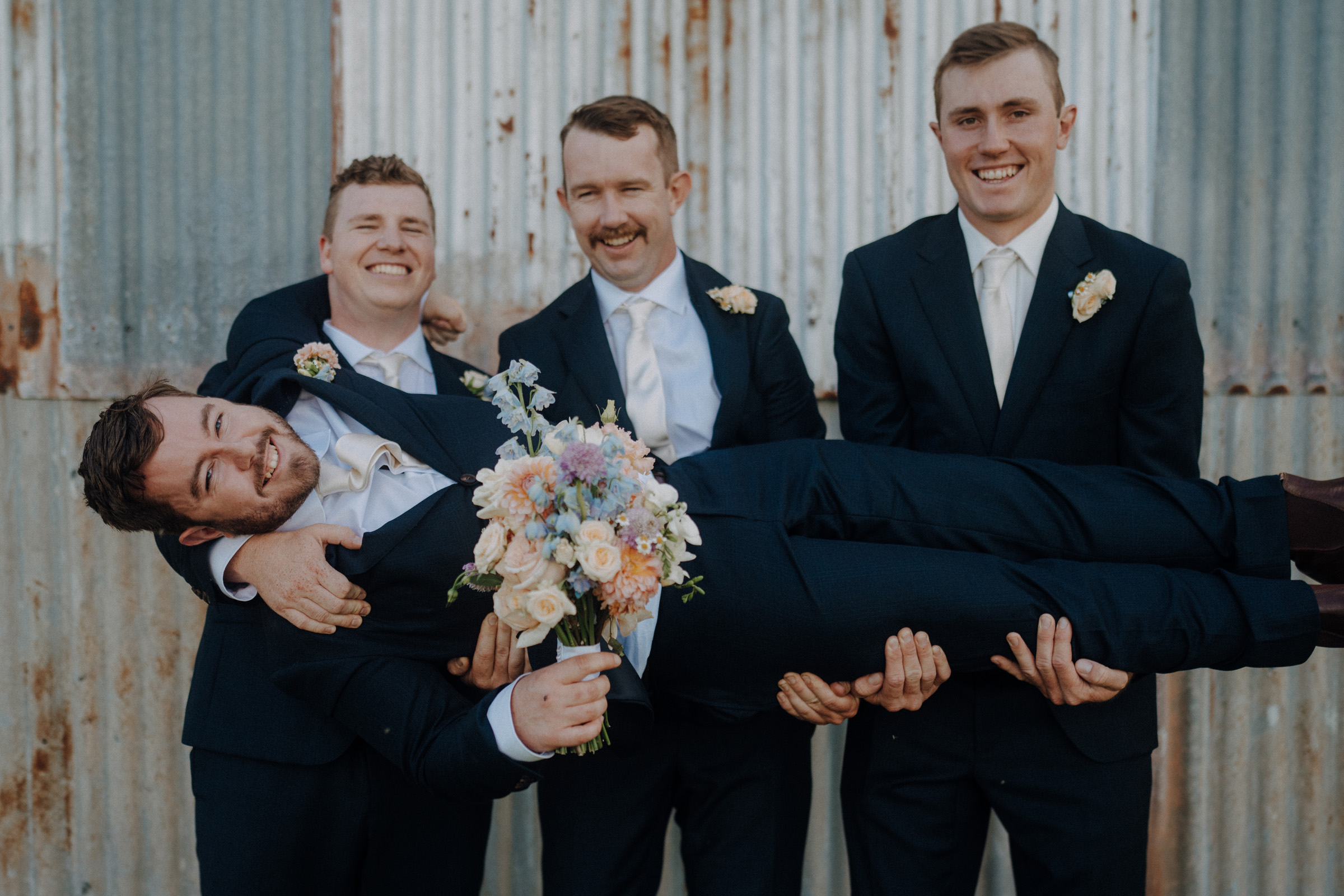 Four men in suits hold another man horizontally. The man being held holds a bouquet. They are smiling in front of a corrugated metal wall.