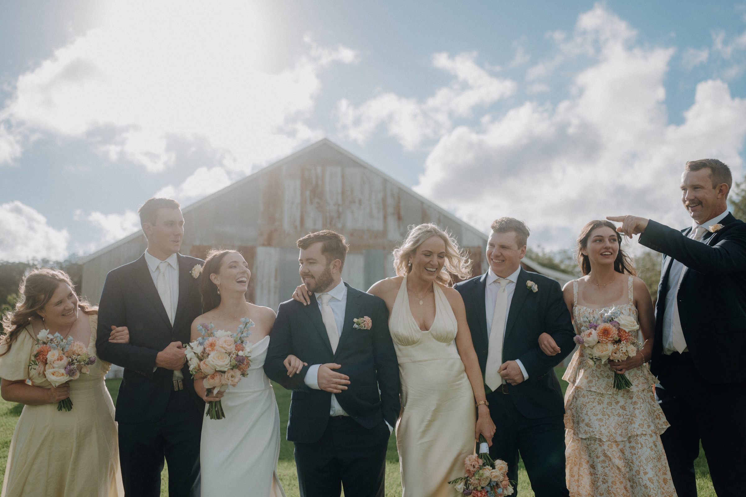 A wedding party stands outdoors, smiling and holding bouquets, with a barn in the background under a cloudy sky.