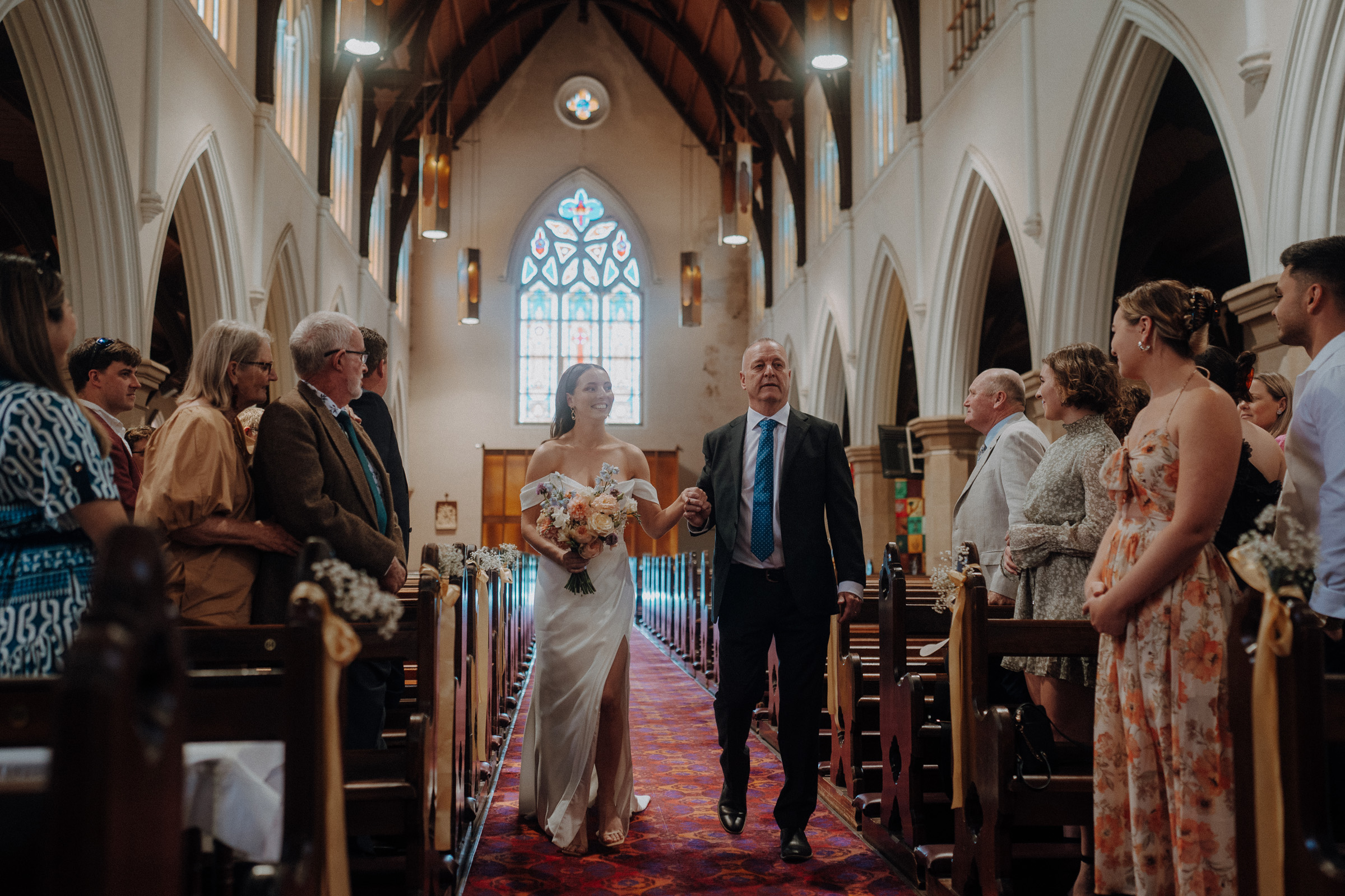 A bride and an older man walk down the aisle of a church, lined with guests on both sides. The bride is holding a bouquet, and the church features large arched ceilings and a stained glass window.