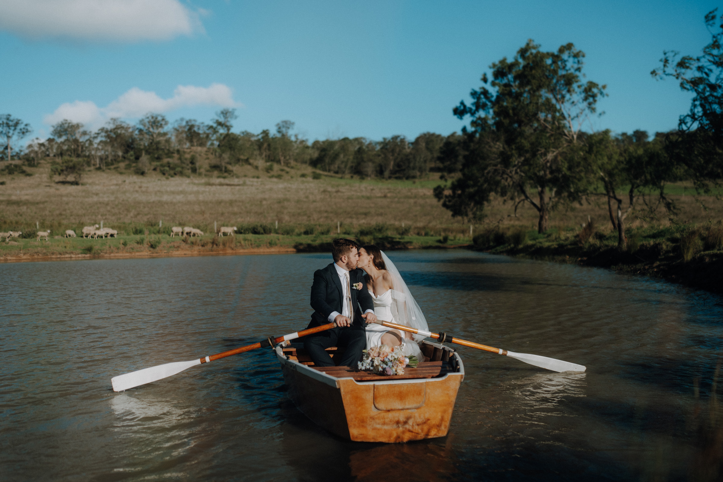 A bride and groom share a kiss while sitting in a wooden rowboat on a calm lake, surrounded by lush greenery under a clear blue sky.