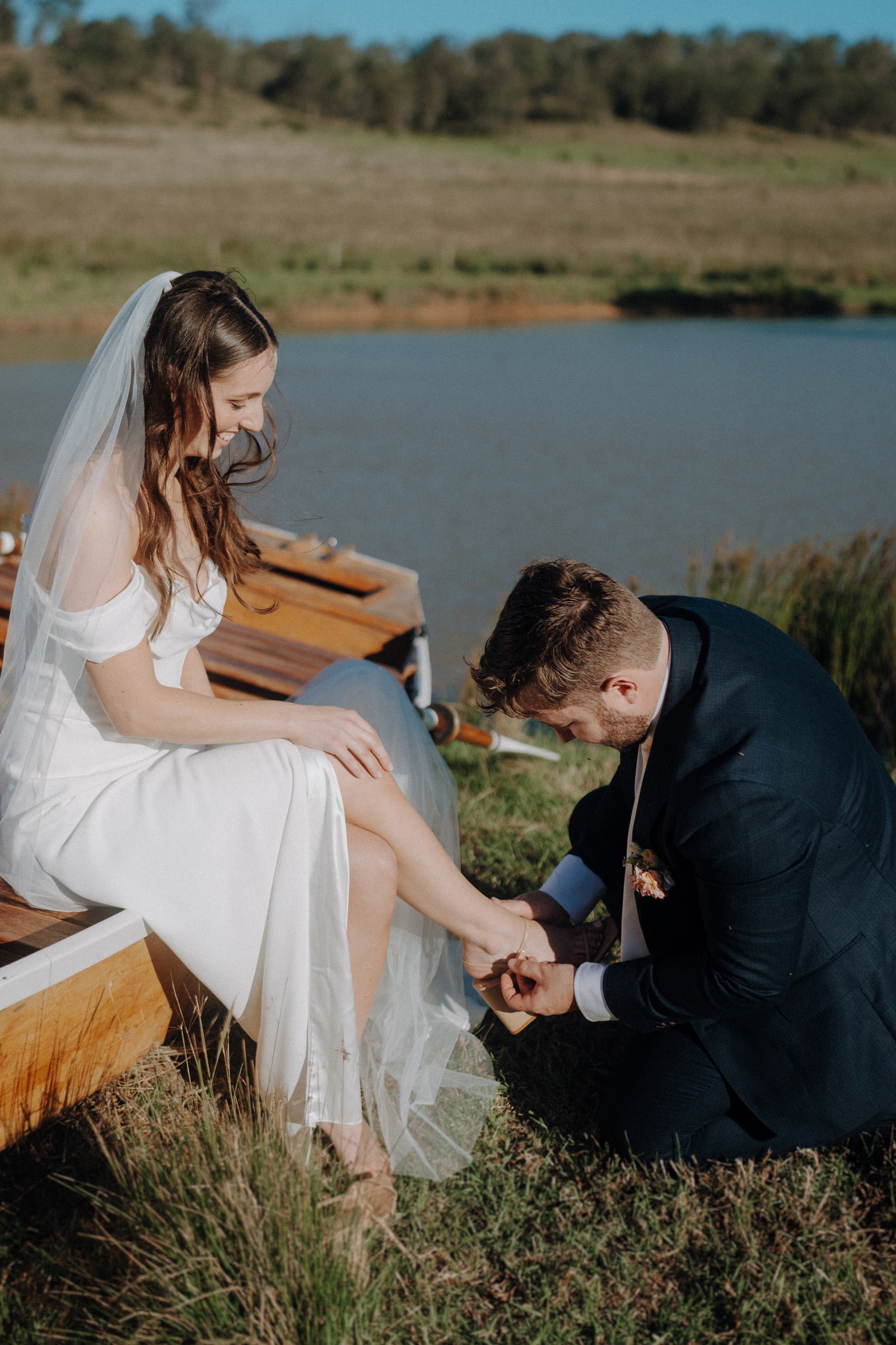 A bride sits on a wooden boat by a lake, while a groom kneels, adjusting her shoe. They are outdoors on a sunny day with grassy surroundings.