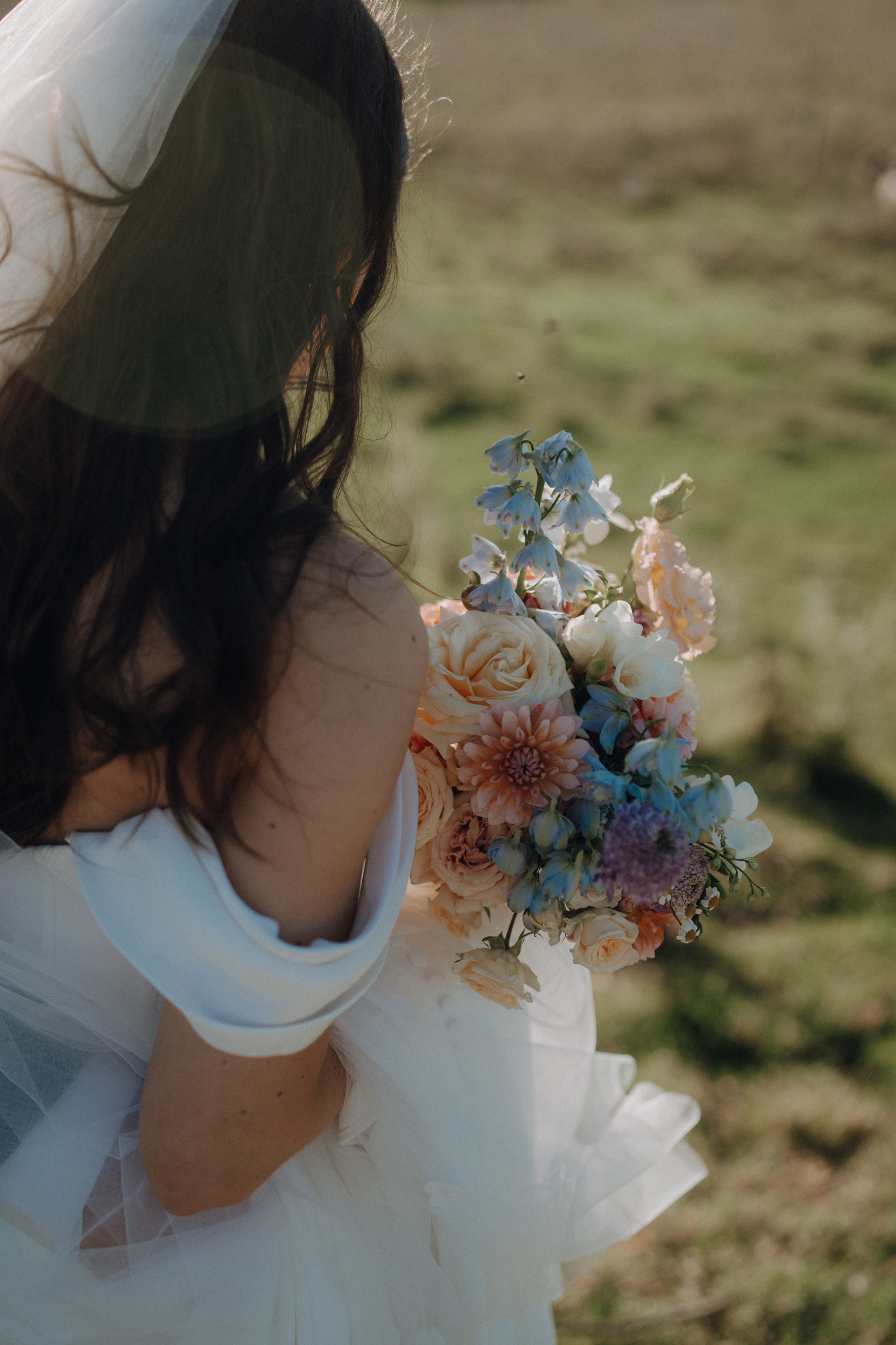 Bride in white dress holding a bouquet of roses, dahlias, and blue flowers, standing on grass with her back partially turned.