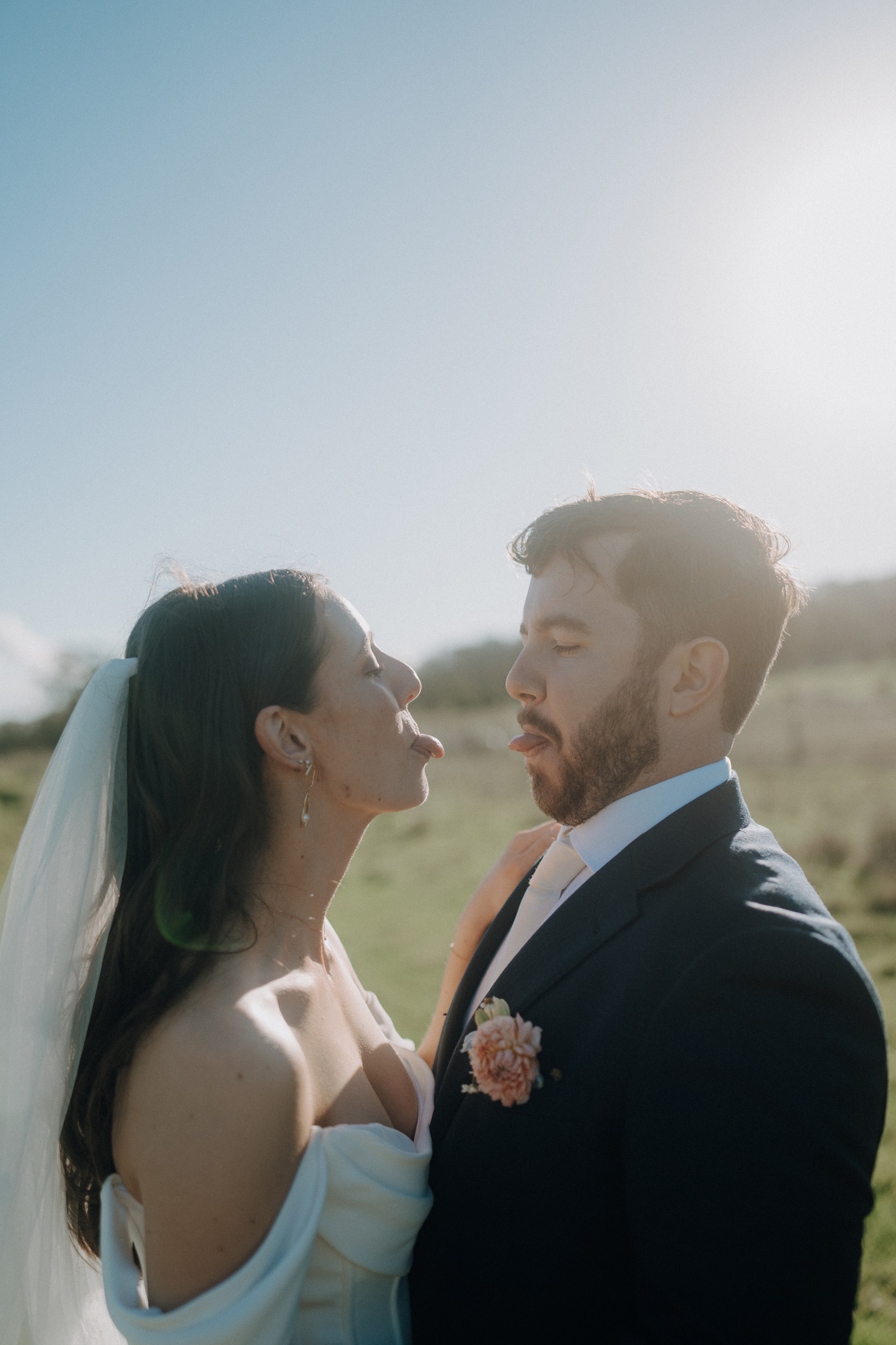 A bride playfully sticks out her tongue at a groom outdoors on a sunny day, with greenery in the background.