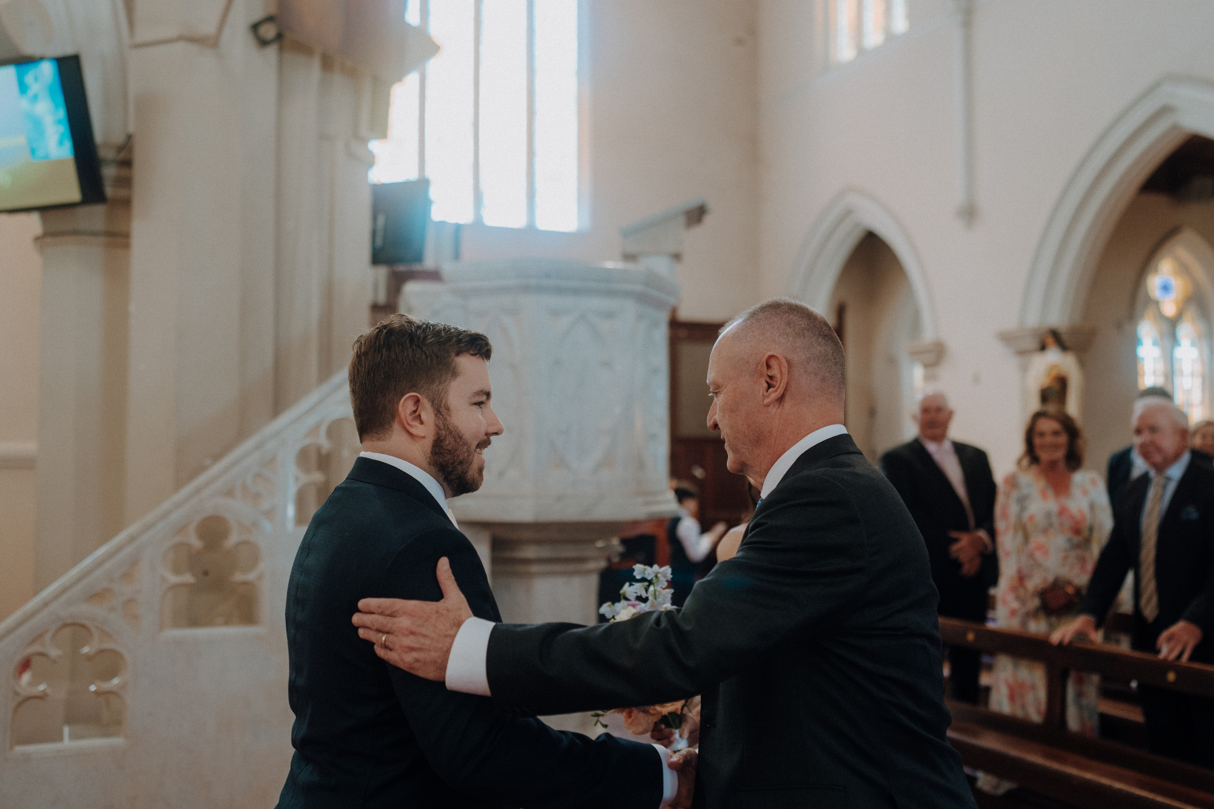 A man in a suit greets another man with a handshake inside a church, as others look on in the background.