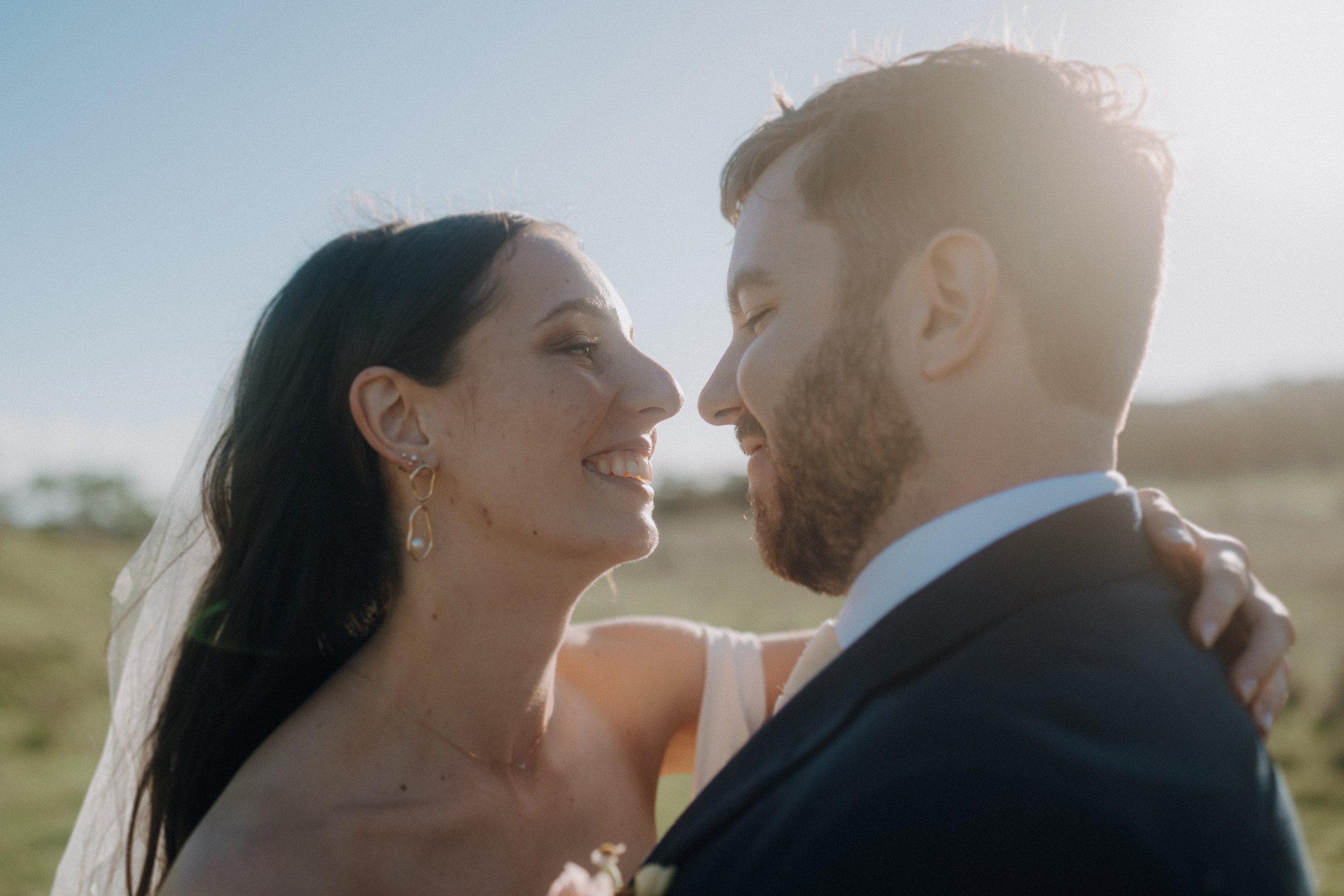 A bride and groom embrace outdoors, smiling at each other under clear skies. She wears a veil; he wears a suit.